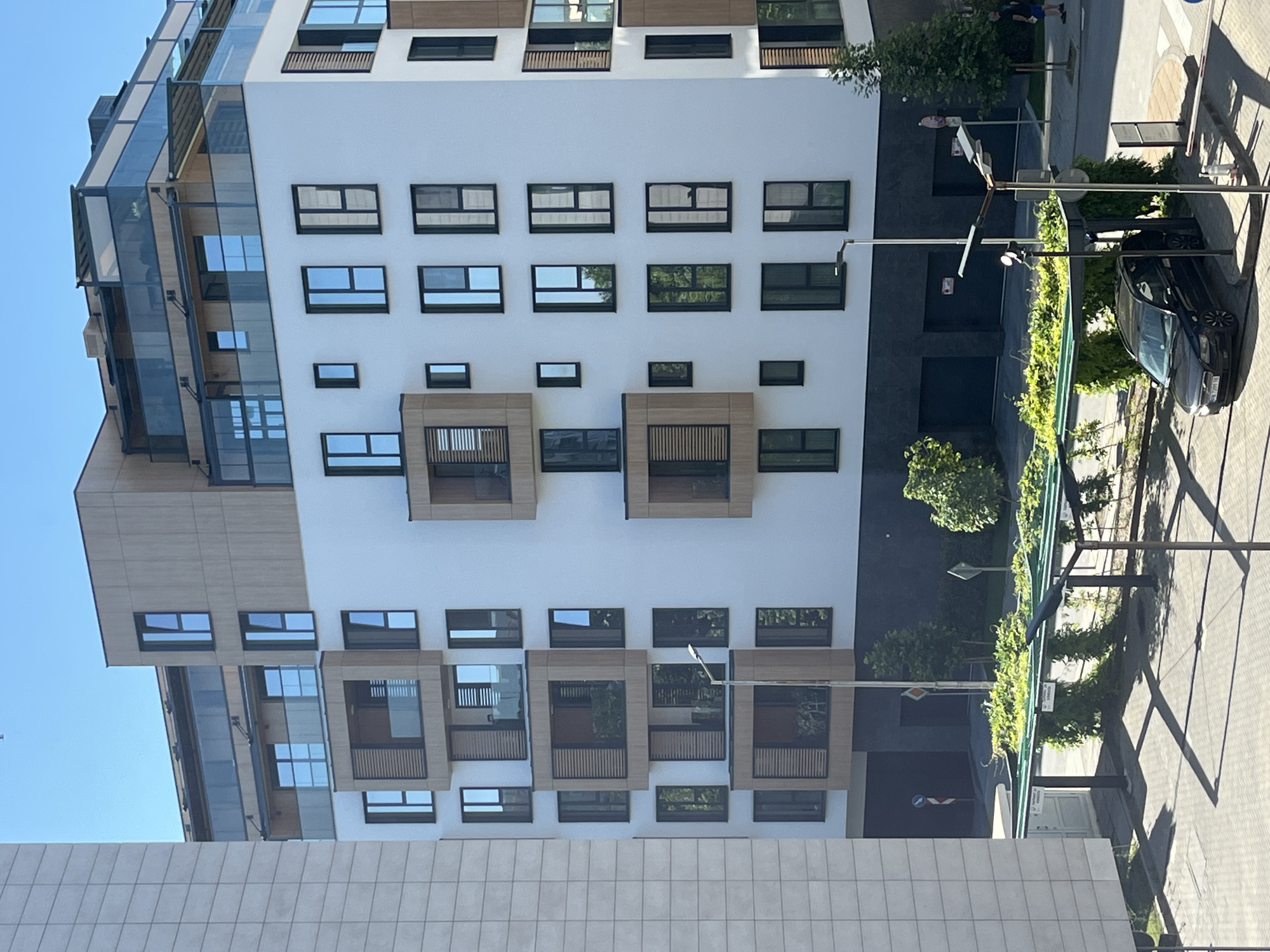 The image captures a modern residential building with multiple balconies and large windows. The building's facade is a combination of white plaster and wooden accents, with some balconies featuring glass railings and wooden shutters. A car is parked under a covered structure with lush greenery growing over it. The surrounding environment appears to be urban, with paved ground and trees. The sky is clear and blue, indicating a sunny day. The architecture suggests a contemporary design. There are no visible people engaged in any specific activity in this frame. The car is black and appears to be a sedan. The building has several stories, with the upper floors featuring more expansive balconies and glass enclosures. A lamp post is visible in the foreground. A portion of another building with a textured facade is visible to the left of the main subject.