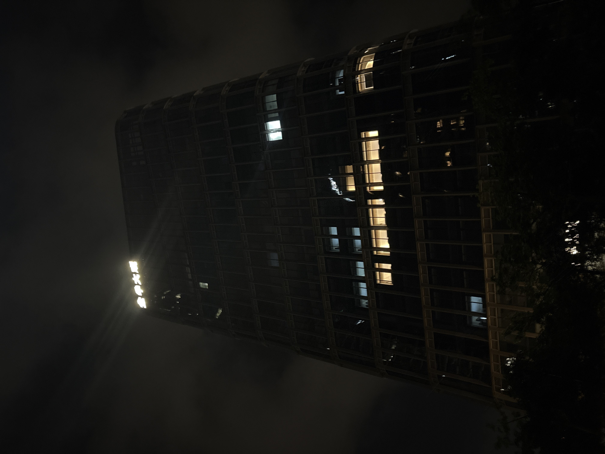 A nighttime, low-angle shot depicts the tall, dark facade of the Guangzhou Bank Tower in Haizhu District, China. The building's sleek, curved design is emphasized by the dark sky and minimal exterior lighting, except for illuminated signs at the very top which read "广州银行" (Guangzhou Bank). Several windows are lit from within, casting warm yellow and white light onto the dark glass, with one section on a lower floor showing a more intensely lit, expansive interior space. The base of the building is obscured by the dark silhouettes of trees and foliage. The sky is cloudy, with some areas appearing lighter, possibly due to atmospheric haze or distant city lights.