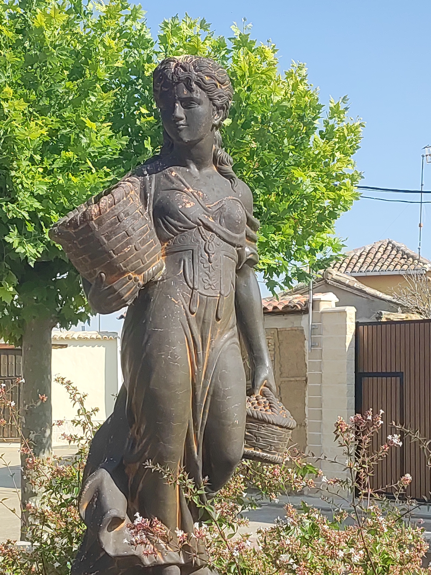 A bronze statue of a woman stands in the foreground, holding a basket in her left arm and another basket filled with fruit in her right hand. She is dressed in a long, flowing garment with intricate detailing on the bodice. The statue is weathered, with some patches of rust. Behind the statue, a large, leafy green tree dominates the upper left portion of the image. To the right of the tree, a section of a building with a tiled roof is visible. The background also includes a wooden fence and a clear blue sky. The statue is located in the town of Autillo de Campos, Spain, and appears to be in a public space, possibly a plaza or park. The lighting suggests it is daytime, with sunlight casting shadows on the statue and foliage. Small flowering plants are growing at the base of the statue, adding a touch of color to the scene.
