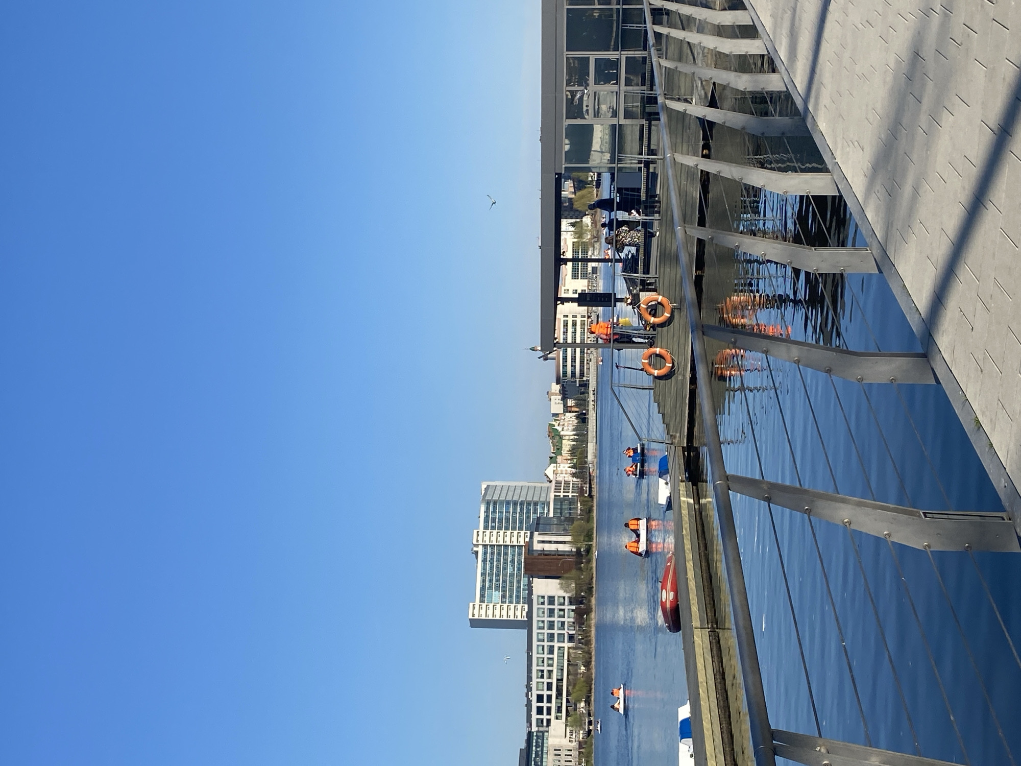 The image depicts a waterfront scene in Kazan, Russia, with a clear blue sky dominating the view. In the water, several paddle boats are visible, each carrying people wearing orange life vests. Buildings of varying heights can be seen along the waterfront, indicating an urban environment. A modern bridge or walkway with metal railings is in the foreground, partly obscuring the view of the water and the city beyond. A bird is flying in the open sky. Safety buoys are located on the side of the bridge or dock.