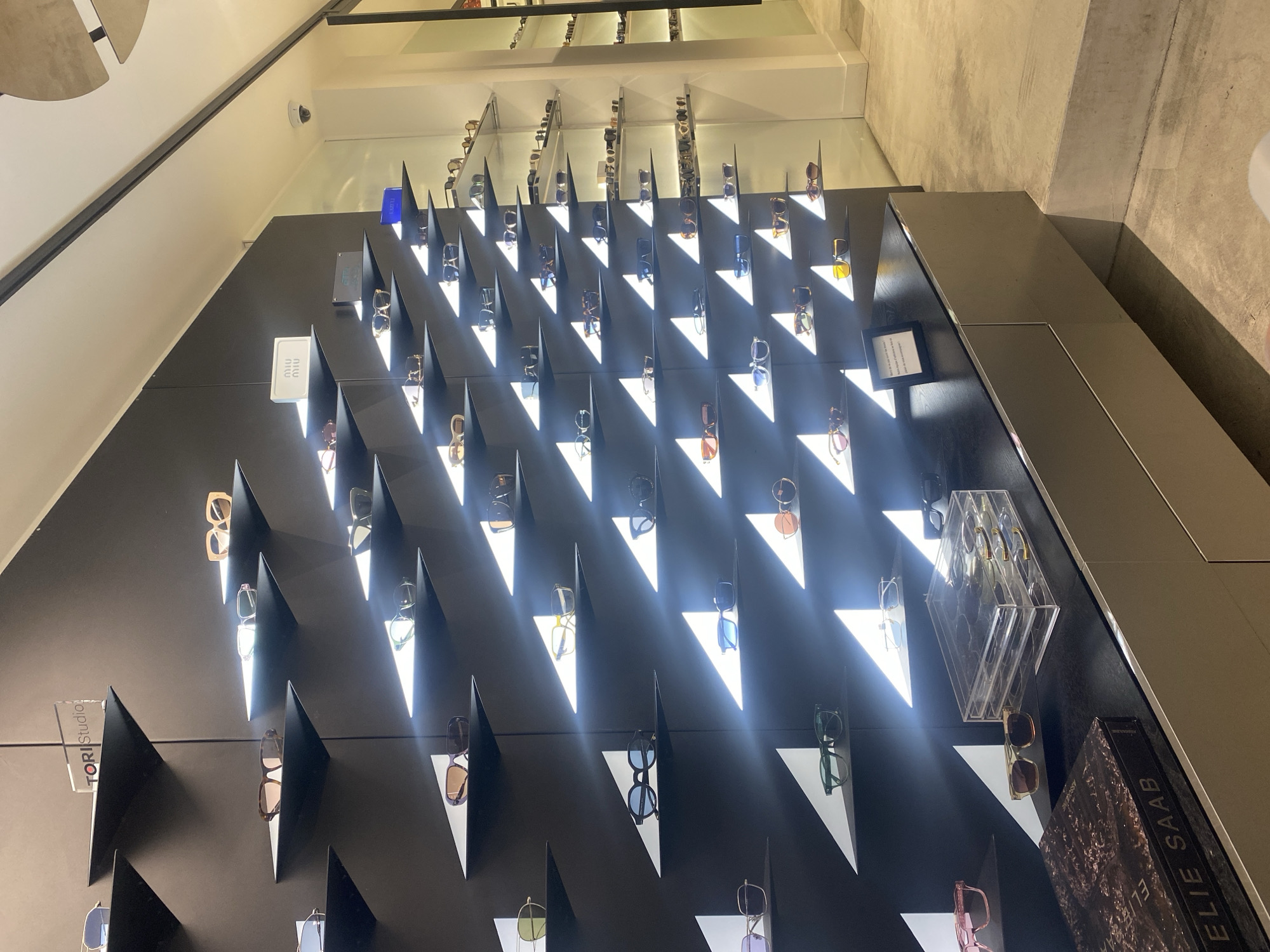 The image depicts a display of sunglasses in what appears to be a retail setting. The sunglasses are arranged on a black wall with triangular shelves, illuminated from behind to create a dramatic lighting effect. The display features various brands, including "TORI Studio", "MIU MIU", and "ELIE SAAB", as indicated by branding labels and signage. A selection of sunglasses is also visible on shelving above the displayed wall. Some sunglasses are arranged in a clear acrylic display case on the right side of the wall. Based on the presence of French branding and luxury products, the location may likely be a retail establishment located in Paris, France, but no further elements indicating Parisian ambiance are present in the image. No people are visible in the photograph.