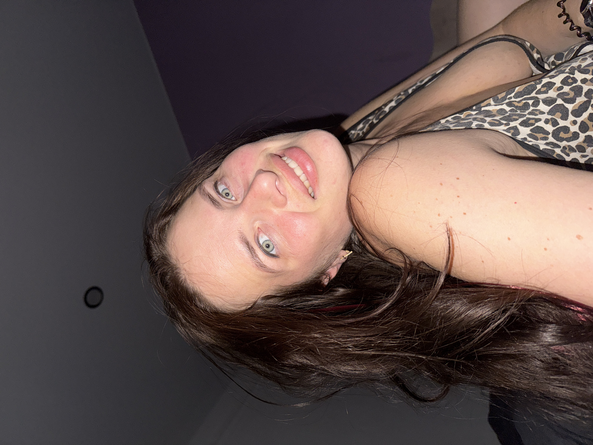 A woman with long, dark brown hair and light blue eyes smiles at the camera. She is wearing a leopard print top and is in a dimly lit room. Her head is tilted back, and her hair is spread around her. The background is dark, with a hint of purple on the right side. There is a circular object on the ceiling in the upper left corner of the image.