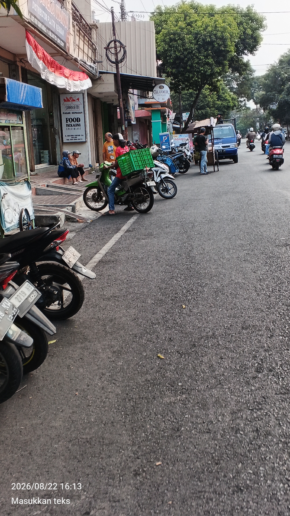 The image depicts a street scene in Malang, Indonesia. In the foreground, several motorcycles are parked on the side of the road. A green crate is affixed to the back of a green motorcycle, suggesting a delivery or transportation task. A person wearing a red shirt and blue jeans is seated on this motorcycle, holding onto the crate.
On the left side of the street, a building displays a prominent Indonesian flag. The building appears to be a commercial establishment with signage indicating "SARLEM" and "FOOD PACKAGING SPECIALIST." Several individuals are seated on steps in front of the building, observing the street.
Further down the street, other motorcycles and a blue van are visible. Trees line the side of the road, and the pavement shows signs of wear. The overall impression is of a busy street with commercial activity and transportation. The timestamp "2026/08/22 16:13" is visible in the bottom left corner, along with the text "Masukkan teks."