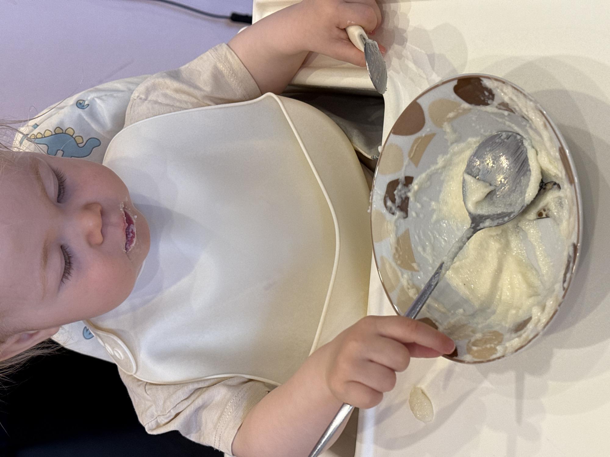 A top-down shot shows a child with fair skin and closed eyes, wearing a beige long-sleeved shirt and a light-colored bib with a dinosaur pattern, sitting in a high chair. A beige silicone bib covers most of the child's torso. The child's face is tilted slightly to the left, and there appears to be food residue on their lower lip.
To the right, a round plate with a brown and beige patterned rim contains the remnants of a creamy substance, possibly food, with a metal spoon resting on top. A metal utensil, possibly a knife or spoon, is visible near the upper right edge of the plate, held in the child's hand. Another metal spoon is visible, being held in the child's hand in the lower left of the plate.
A blob of food has spilled from the plate onto the white table surface.