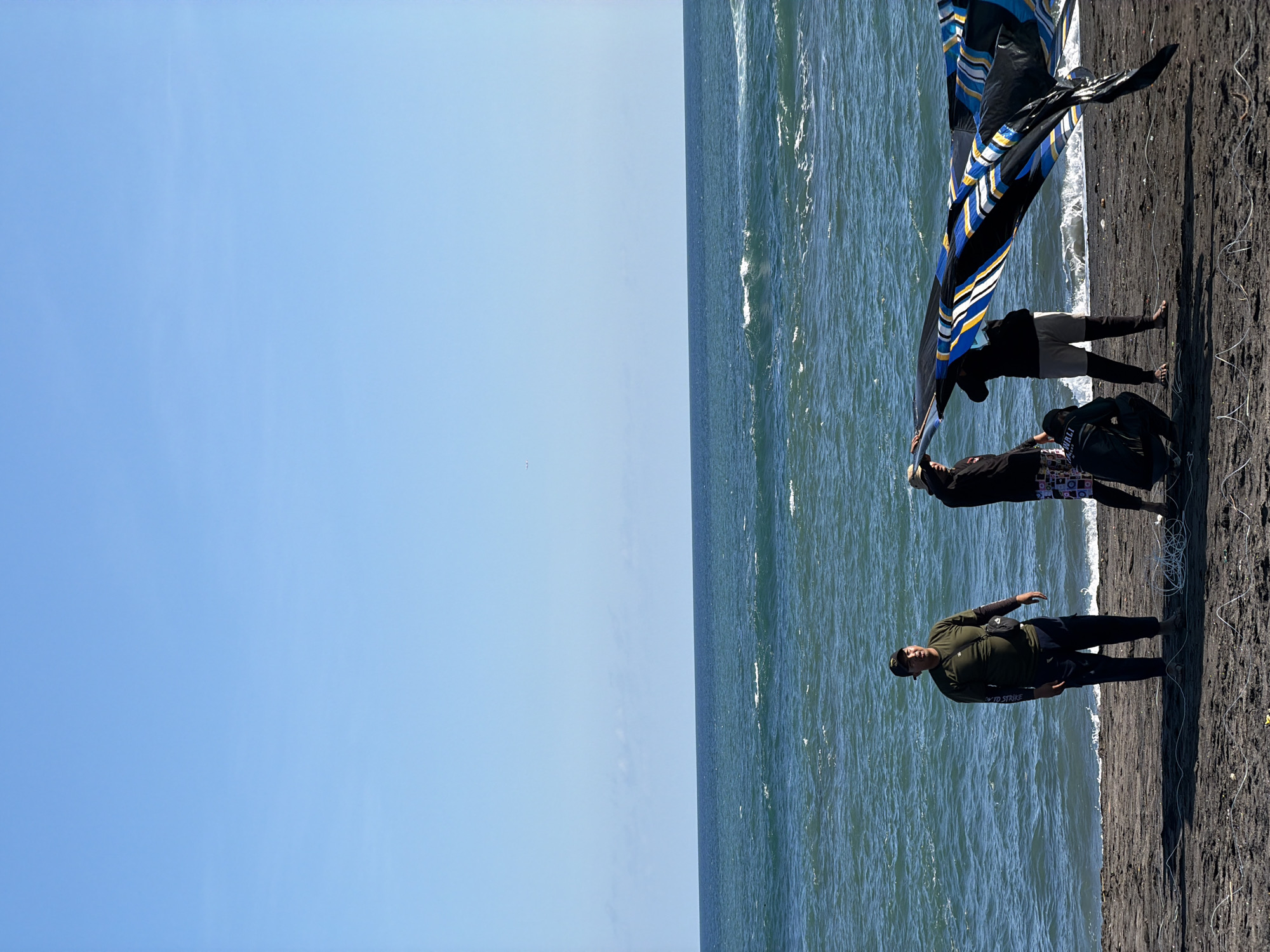 On a sunny day, at a beach in Mengwi, Indonesia, three people stand on dark sand next to the ocean. The sky is a clear blue, and the water is moderately choppy. Two people are holding a large kite-like object with blue, black, and white stripes. One person is looking down. The third person, who is wearing a green shirt, is standing to the side with their arms outstretched.