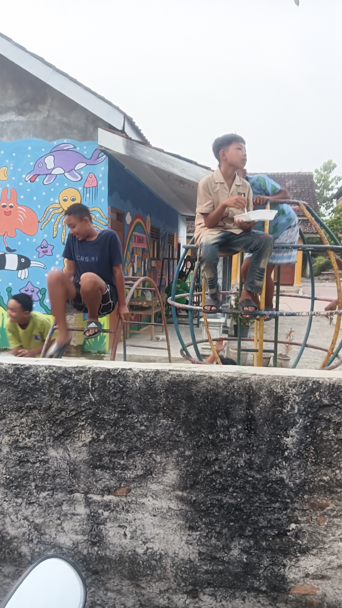 The image captures a scene at what appears to be a playground with a mural-decorated building in Tanggan, Indonesia. In the foreground, a textured, grey concrete wall dominates the lower portion of the frame. Several children are visible on and around a circular metal playground structure. One boy in a light brown shirt and blue jeans is seated on the structure, holding a white plate. Another boy in a dark blue t-shirt and checkered shorts is in motion, appearing to jump or climb on the playground equipment. To the left, a child in a yellow shirt is lying down. The background features a building with a blue wall adorned with a colorful mural depicting marine life and other playful imagery. The sky is overcast.