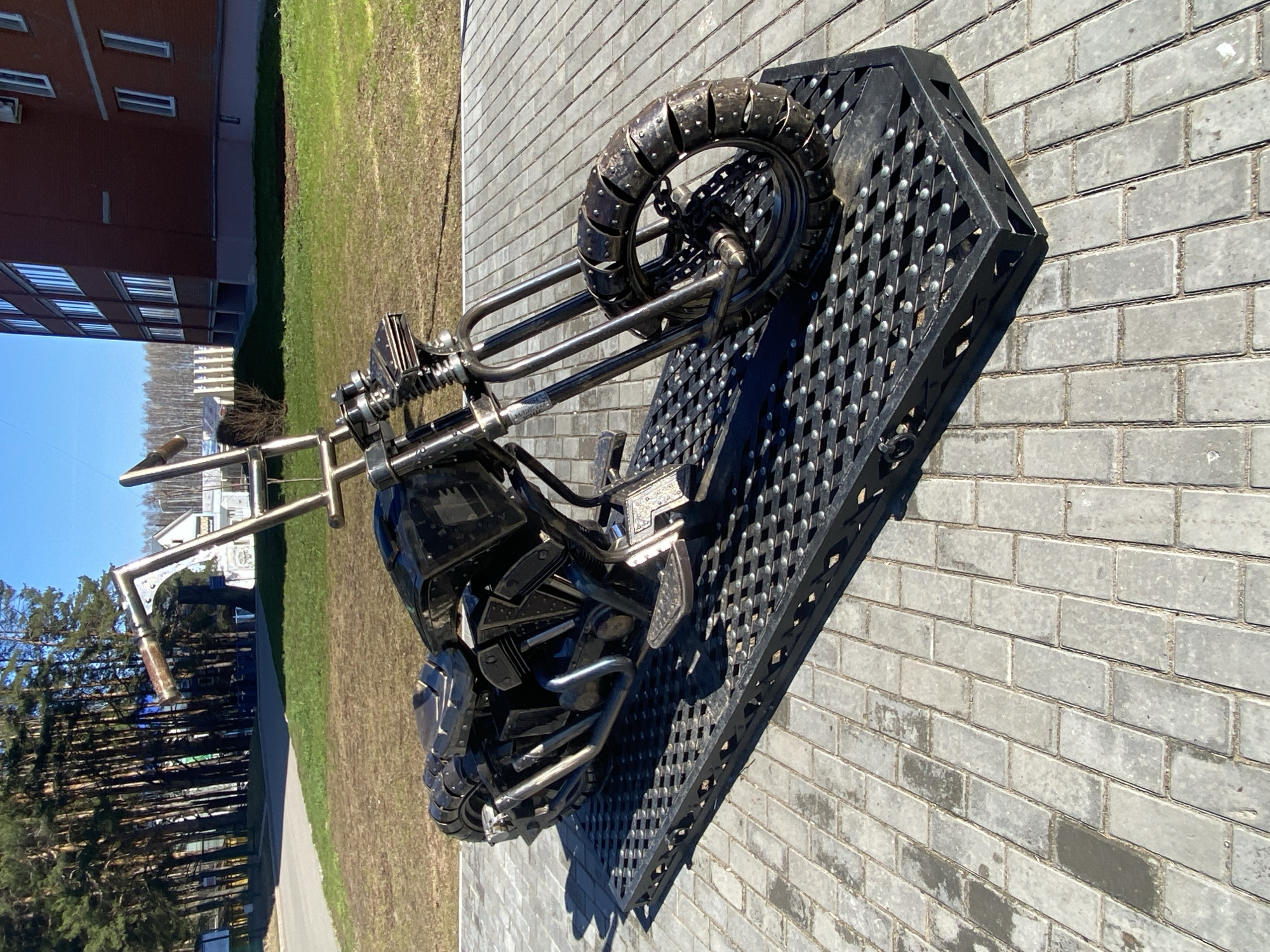 A metal sculpture of a motorcycle rests on a metal grate, placed on a gray brick surface. The sculpture is detailed, with visible engine components, handlebars, and wheels with studded tires. In the background, a building with multiple windows sits adjacent to a green lawn. Further back, a row of trees lines a pathway. The scene is set in Chaikovsky, Russia.