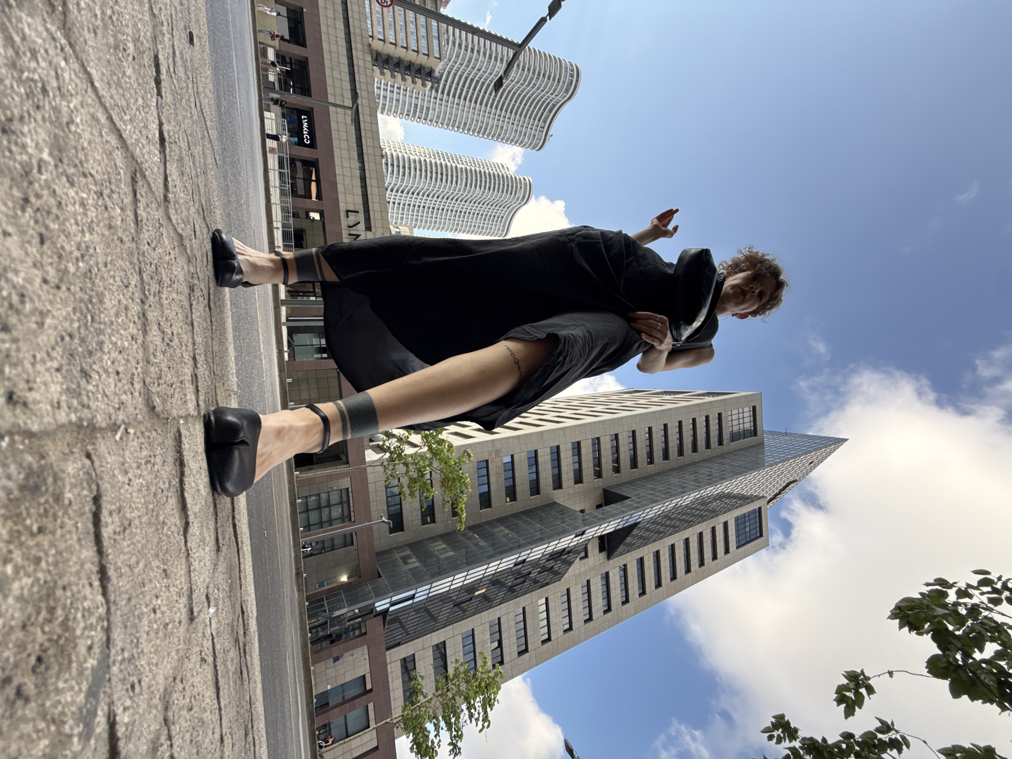 A low-angle shot captures a person standing on a sidewalk in Tel Aviv, Israel. The individual is wearing a black dress, black split-toe shoes, and has distinctive black tattoos on their ankles and calves. They are holding a black bag on their shoulder.
In the background, several modern buildings are visible. To the left, two white, wavy high-rise apartment buildings stand against a blue sky with scattered clouds. To the right, a tall, angular gray skyscraper with reflective windows dominates the skyline. In front of the skyscraper, a lower, brown building with large windows and illuminated signage, including "LYNK & CO," is visible. Trees with green leaves are planted near the skyscraper. The pavement in the foreground is made of gray concrete slabs. The scene appears to be a street in an urban environment.