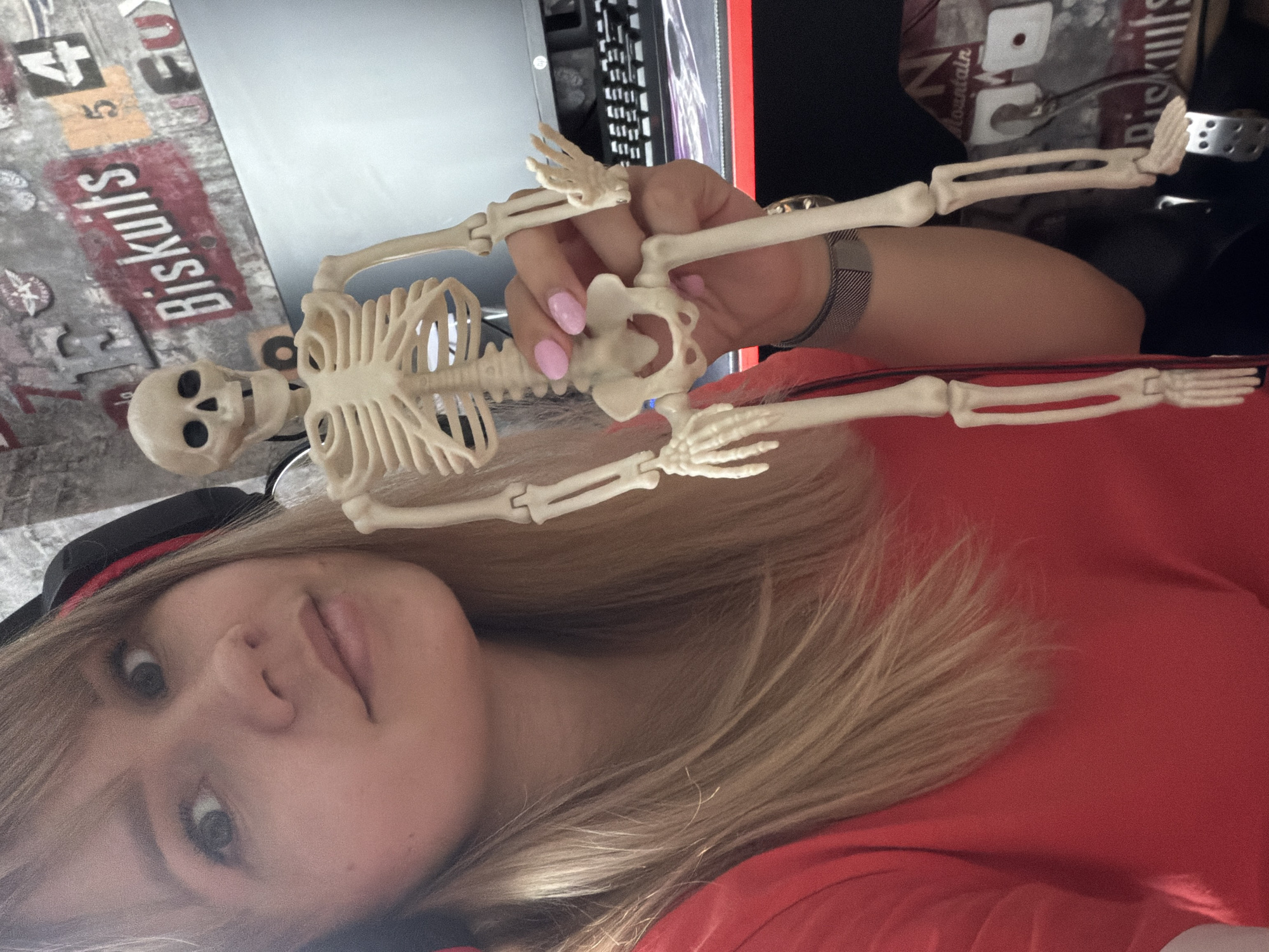 A person with blonde hair and a black cap is holding a toy skeleton. The person is wearing a red shirt and has a wristwatch. The background includes a computer, keyboard, and a wall with "Biscuits" written on it. The location is Unknown, Unknown.