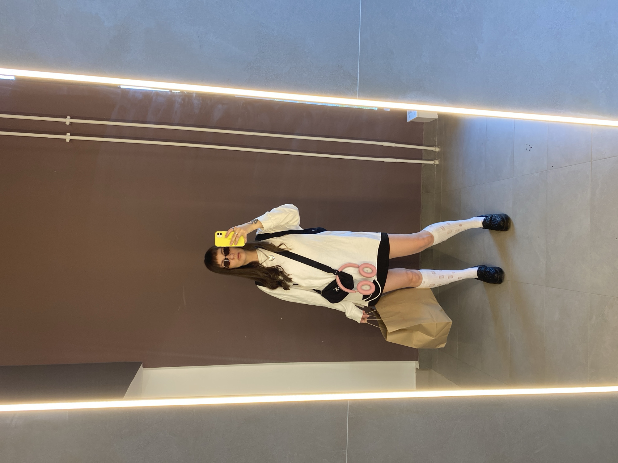 A person stands in a corridor with brown walls and gray tiled floors in Izhevsk, Russia. The person is wearing a white long-sleeve shirt over a black skirt, knee-high white socks, and black shoes. They are holding a yellow smartphone in their right hand, taking a selfie. A black crossbody bag and pink headphones are worn over their left shoulder, and they are holding a brown shopping bag in their left hand. Fluorescent light bars illuminate the corridor's ceiling. White pipes are visible along the brown wall.