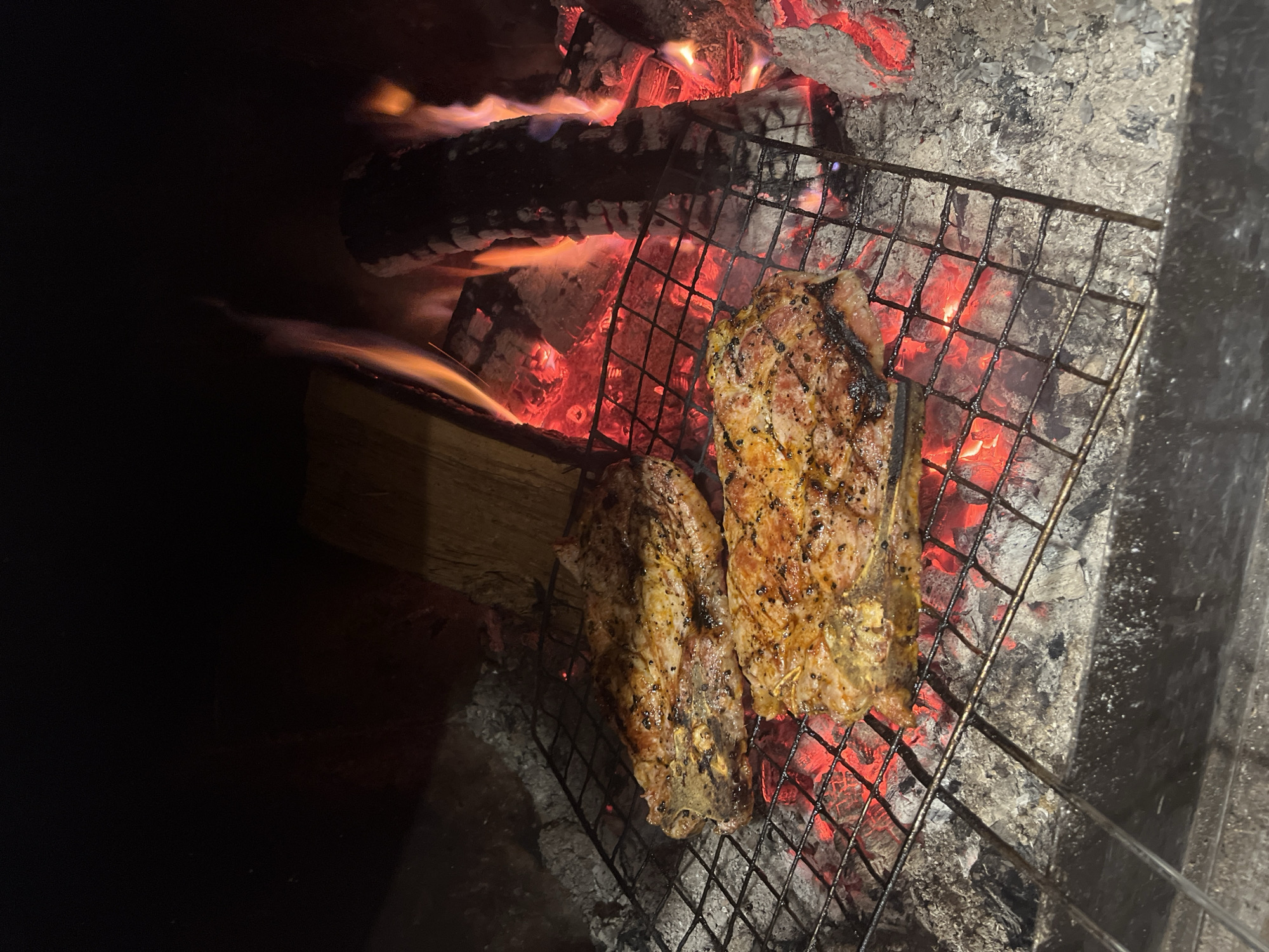 The image depicts two pieces of meat grilling on a metal grate over a fire. The fire consists of burning wood and embers. The grate is positioned over the fire, with the meat placed directly on the grate. The meat is visibly seared and appears to be seasoned. The background is dark, suggesting the grilling is taking place at night or in a dimly lit area. The scene is located outdoors, judging from the embers of the fire. No information regarding the city of Varshilo, Bulgaria, can be extracted from the image.