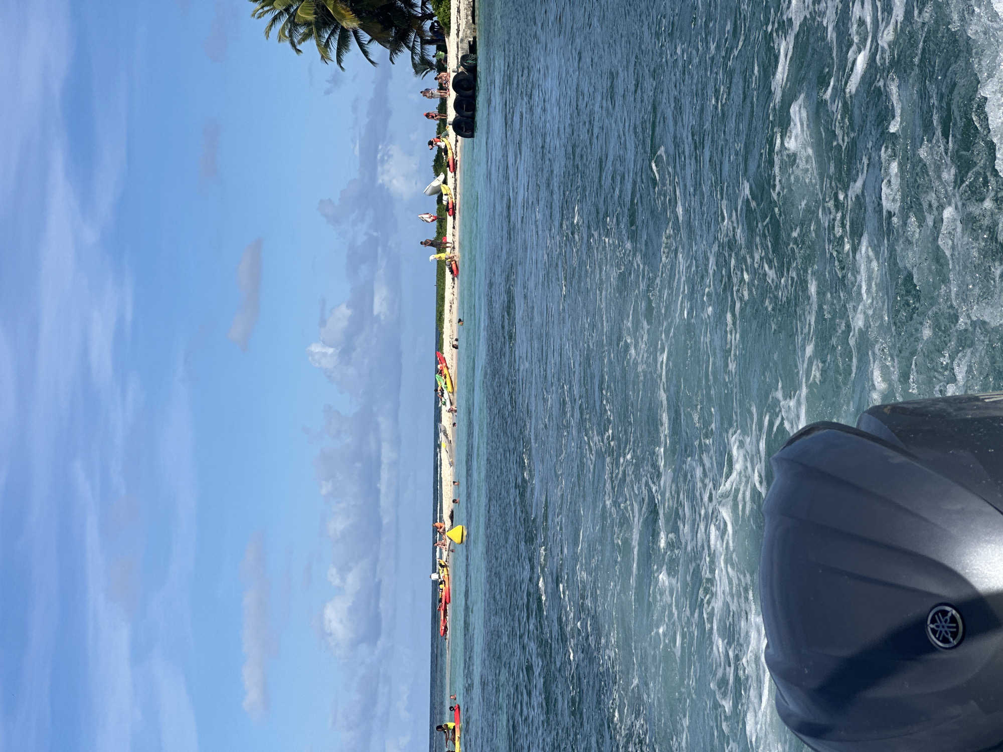 The image, possibly taken from a boat, shows a beach scene in Le Gosier, France. The left side of the image features a blue sky with scattered clouds, gradually transitioning to a sandy beach lined with palm trees and several individuals. Many colorful kayaks rest on the sand. The right side of the image is dominated by blue water with visible wakes, suggesting movement of the boat. A motor with a Yamaha logo is visible in the lower right corner. The people on the beach are spread out, some appearing to be near the water's edge. There appear to be tires or dark objects stacked near the far end of the beach.