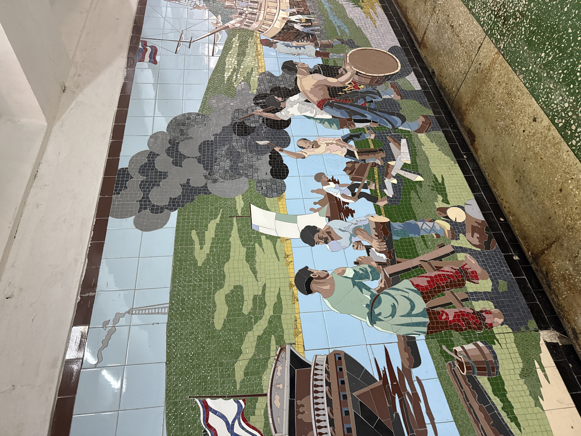 The image shows a mosaic depicting a historical scene, possibly a battle or uprising. The mosaic is located on a wall in Rostov-on-Don, Russia. The scene is composed of various figures in action: some are fighting, others are carrying drums, and some are near ships flying flags. The environment includes land, sky, and ships. Smoke is depicted as rising, likely from cannons. The artwork is rendered in a mosaic style with various colored tiles forming the figures and landscape.