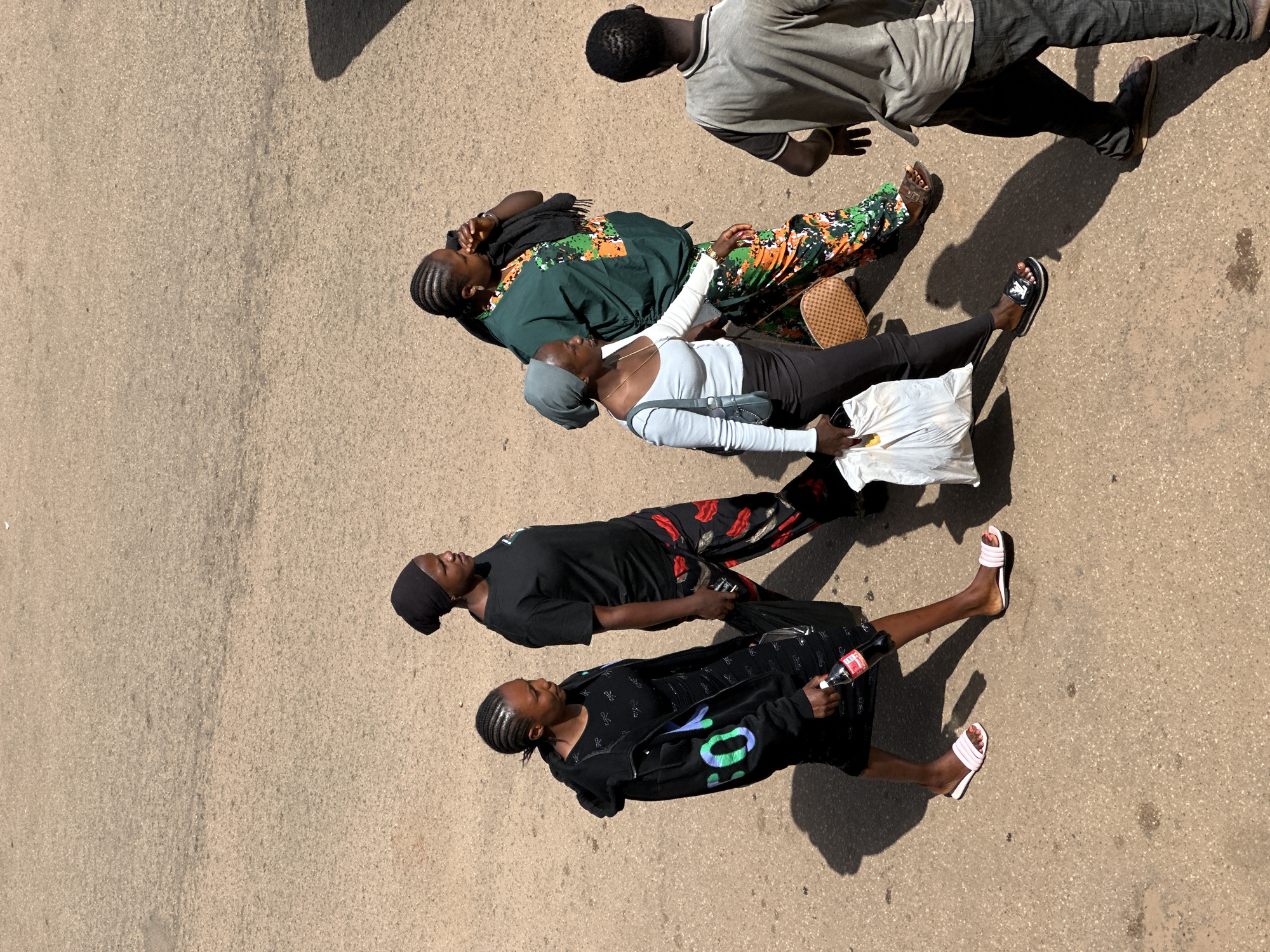In the city of Bukuru, Nigeria, five individuals are walking across a dusty, asphalt surface. The scene is captured from an overhead perspective, with the sun casting distinct shadows.
From left to right:
The first person is a woman wearing a black hooded jacket over a black dress and pink slide sandals. She is carrying a bottle of Coca-Cola in her right hand. Her hair is styled in braids, and she wears a black head covering.
Next to her, a woman in a black t-shirt and patterned pants walks with her head turned to the right. She also wears a black head covering and carries a dark bag in her left hand.
The third person is a woman in a light blue long-sleeved top, black pants, and black slide sandals. She has a grey head covering and carries a white plastic bag in her left hand and a woven bag on her right arm. A light blue purse is visible on her left shoulder.
Behind her, a woman in a dark green top with an orange and green pattern walks, wearing a black head covering. Her right hand is near her face, and she has a black scarf draped around her neck. She wears patterned pants and brown slide sandals.
To the far right, a man is seen from the back, wearing a grey t-shirt and dark pants. He has a short haircut and is walking away from the viewer. His right hand has a yellow bracelet.
The environment is an outdoor setting with a predominantly beige and grey asphalt surface, indicative of a street or public walkway. The lighting suggests a bright, sunny day.