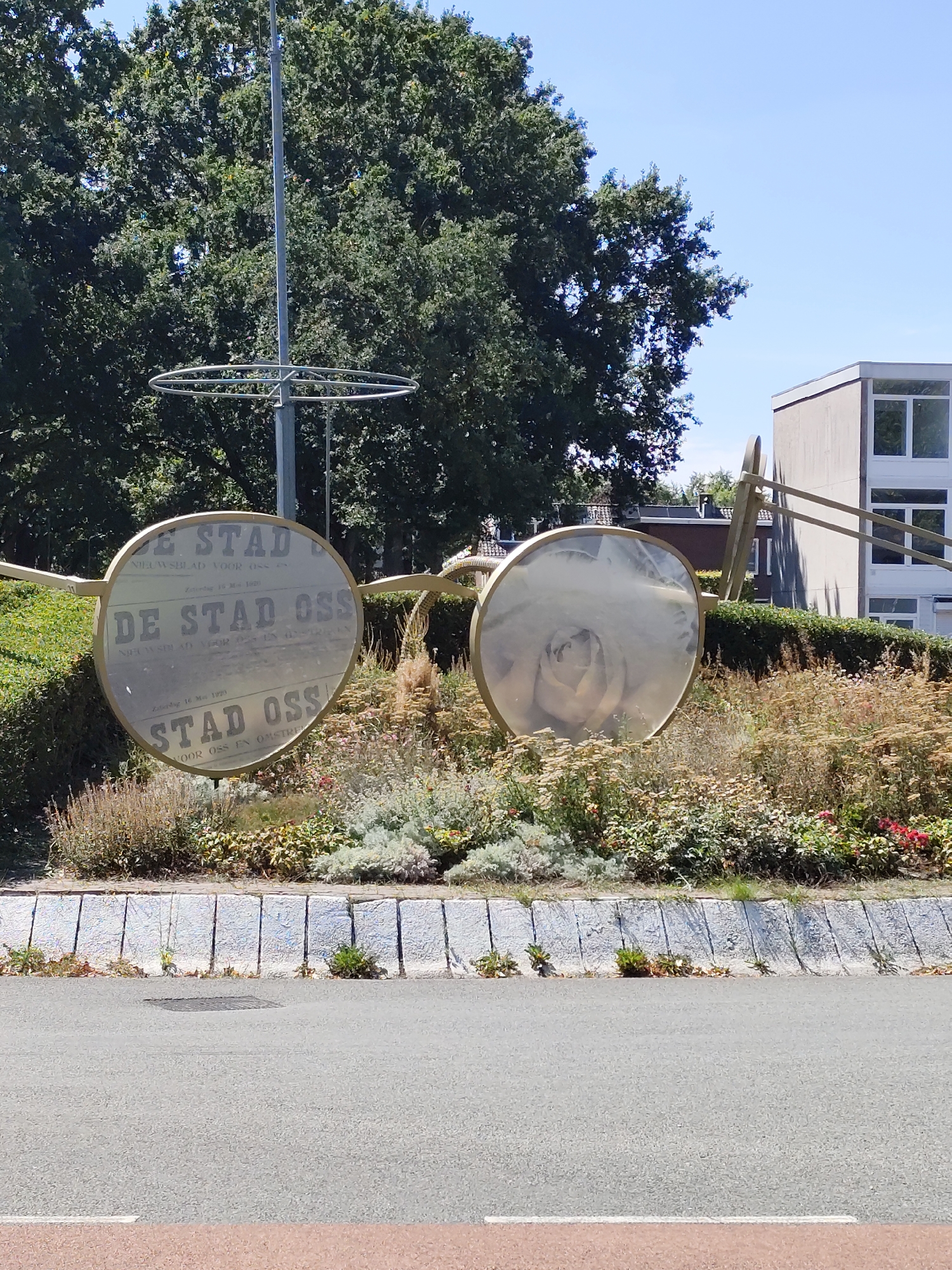The image shows a large outdoor sculpture of eyeglasses, located in the city of Oss, Netherlands. The sculpture is made of a golden-colored material and is positioned in a garden area with various plants and bushes. The lenses of the eyeglasses reflect a newspaper with the headline "DE STAD OSS", which is a local newspaper. In the background, there are trees and a modern building. The ground in front of the garden is a paved road. The sky is clear and blue. There are no people visible in the image.