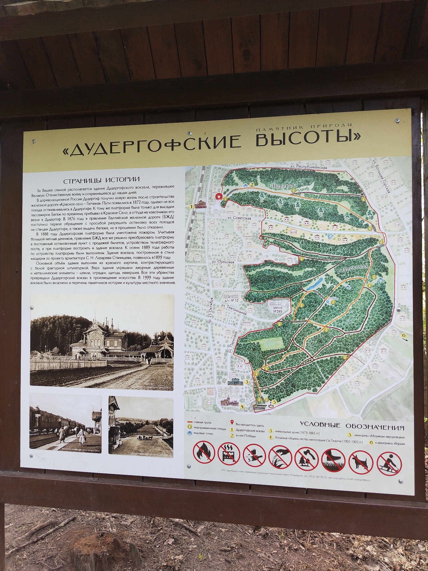Here's a detailed description of the image:
**Scene Overview:**
The image depicts an informational sign or plaque located within the park "Duderhof Heights". The plaque contains a map of the park along with historical information and legends to help visitors understand the park's significance.
**Specific Details:**
* **Plaque Contents:**
* **Top Heading:** The title reads "Duderhof Heights"
* **Map:** A detailed map shows the layout of the park, including paths, and significant landmarks. Numbers on the map correspond to points of interest and informational locations.
* **Historical Text:** A section titled "Pages of History" provides information about the Duderhof train station and its role.
* **Images:** The plaque features historical images of the Duderhof train station and its surrounding area.
* **Legend:** A legend lists the meanings of symbols used on the map.
* **Environment and Context:** The plaque seems to be installed outdoors, likely within the park it describes. Its frame is visible, possibly made of wood.
**Location Clues:**
* The map shows street names that could be used to identify the exact location, such as Republic Street, Soviet Street, and October Street.
* The park is named "Duderhof Heights", which is a place in Saint Petersburg, Russia.
**Additional Information:**
* The informational plaque lists contact details for the Directorate of Specially Protected Natural Areas of Saint Petersburg.