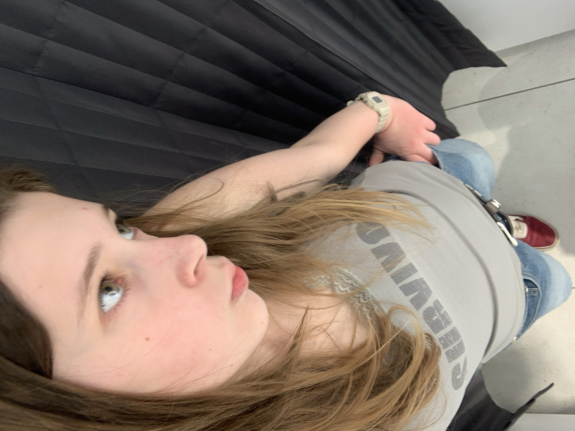 An overhead, slightly angled photograph captures a young person, likely female, reclining indoors. The subject is positioned with her head towards the upper left and her feet towards the lower right of the frame, lying on what appears to be a black, horizontally pleated surface, possibly a bench or padded seating. Her long, light brown hair is spread out around her head and shoulders.
The visible side of her face shows light-colored eyes, possibly green or blue, looking upwards with a neutral expression. She has fair skin and lightly parted lips. She is dressed in a light grey ribbed tank top, which has prominent dark grey text spelling "SURVIVOR" across the chest. She wears light blue jeans and dark red sneakers with white accents. A light-colored digital watch, possibly off-white or beige, is visible on her left wrist.
The background behind her head and upper body consists of the black pleated material. Towards her lower body and feet, a light grey, possibly concrete or tiled, floor is visible. The lighting is soft and even, indicating an indoor setting. The scene conveys a relaxed and casual moment in what could be a public waiting area or similar indoor environment in Belgrade, Serbia.
