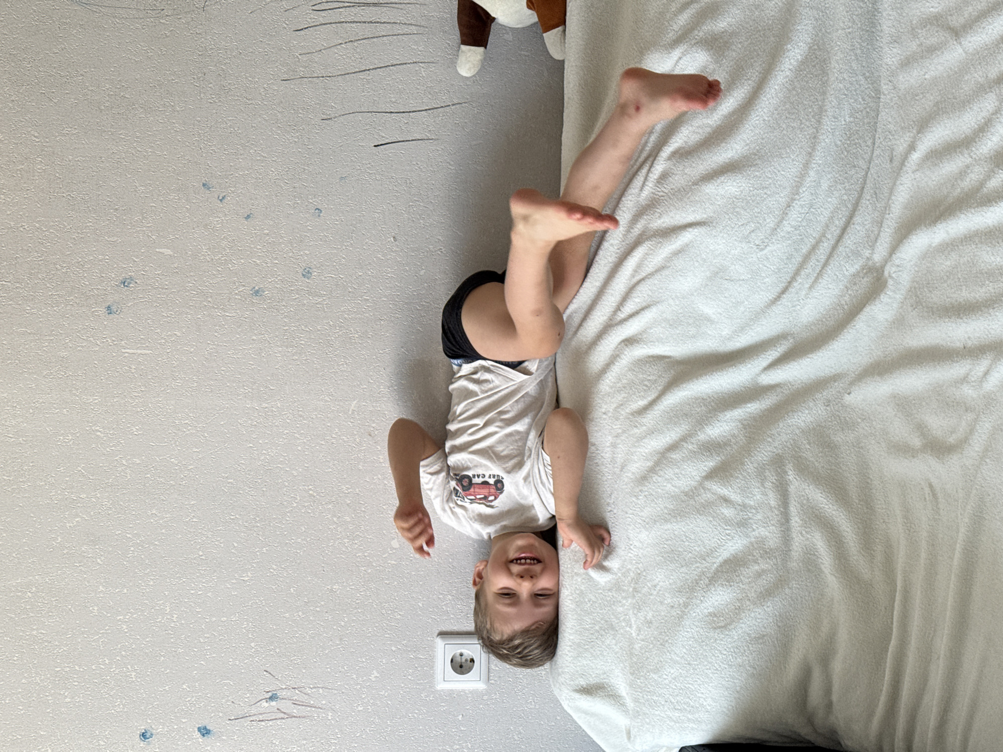 A young boy is lying on his side on a white blanket on what appears to be a bed or sofa. He is wearing a white t-shirt with a red car graphic and black shorts. He is smiling and looking at the camera. His right arm is bent at the elbow, and his left arm is extended behind him. His legs are bent and positioned behind him. The wall behind him is light gray and has some blue smudges and black scribbles on it, suggesting it may have been drawn on by children. A white electrical outlet is visible on the wall to the boy's left. A stuffed animal with brown and white ears is partially visible on the right side of the image. The lighting is bright, indicating it is daytime. The overall scene appears to be in a domestic setting.