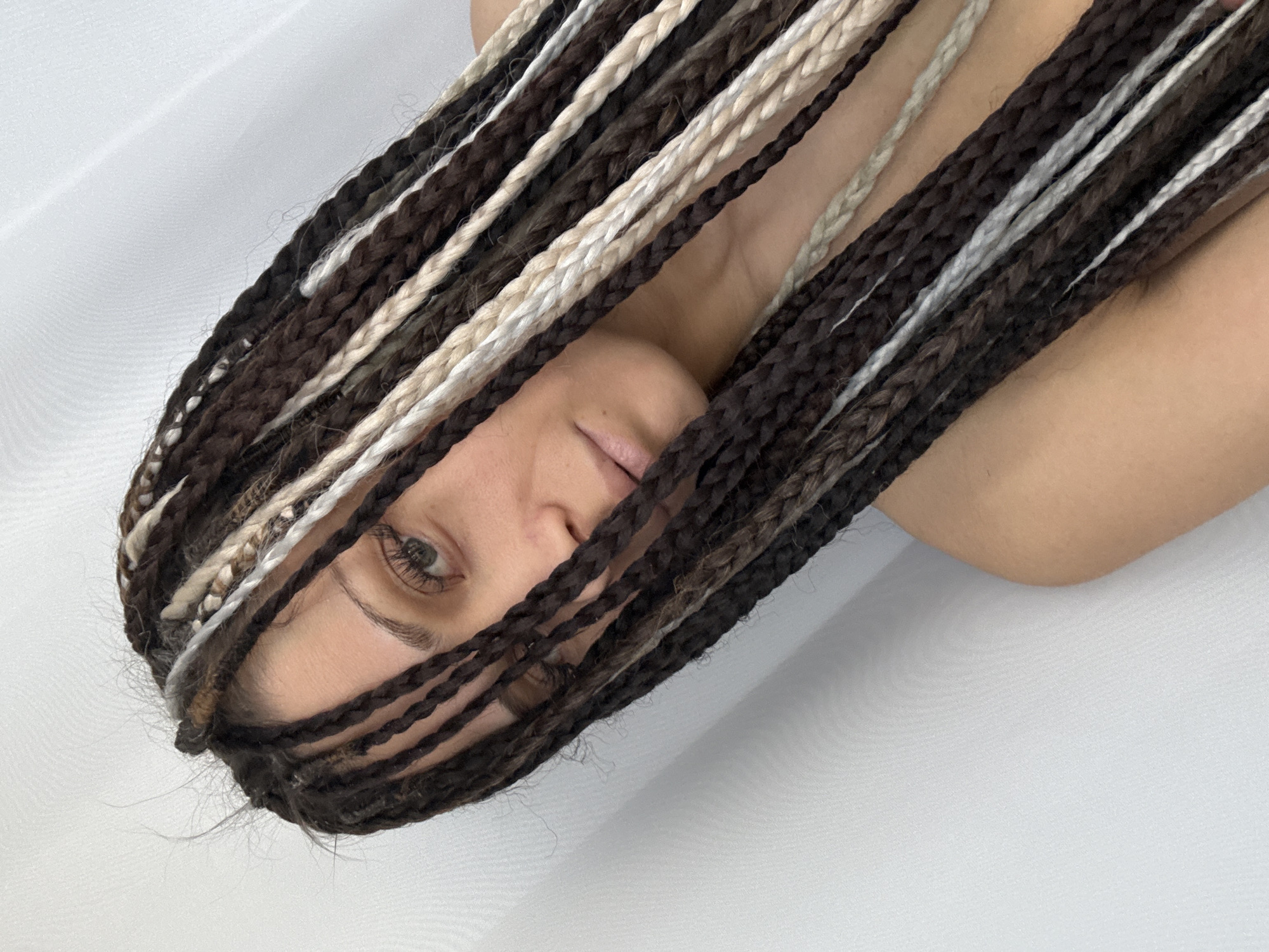 A close-up, angled view shows a person lying on a white surface. The person has a head full of braided hair, in alternating shades of brown and white. The braids partially obscure the face, although one eye and a portion of the nose and mouth are visible. The person's skin is fair. The background is plain white, suggesting a minimalist setting. No specific location is identifiable.