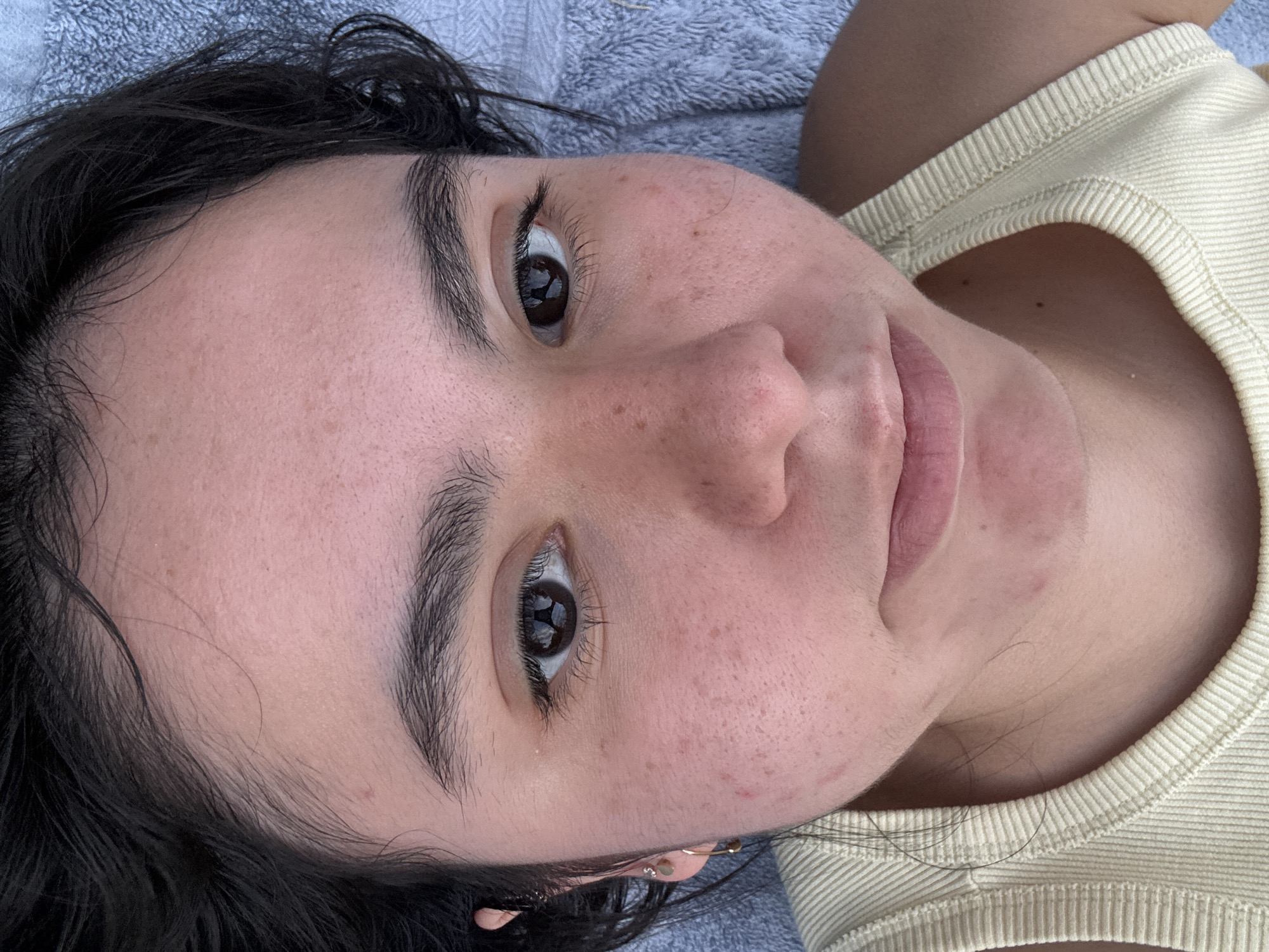 A close-up, tilted portrait shows a person with dark hair, brown eyes, and light skin with freckles lying on a surface, possibly a towel given its texture. They are wearing a light-yellow ribbed tank top. The background is blurred, suggesting a shallow depth of field. The individual is looking upwards towards the camera. There is no indication of the location being in Austin, Texas or any other specific city.