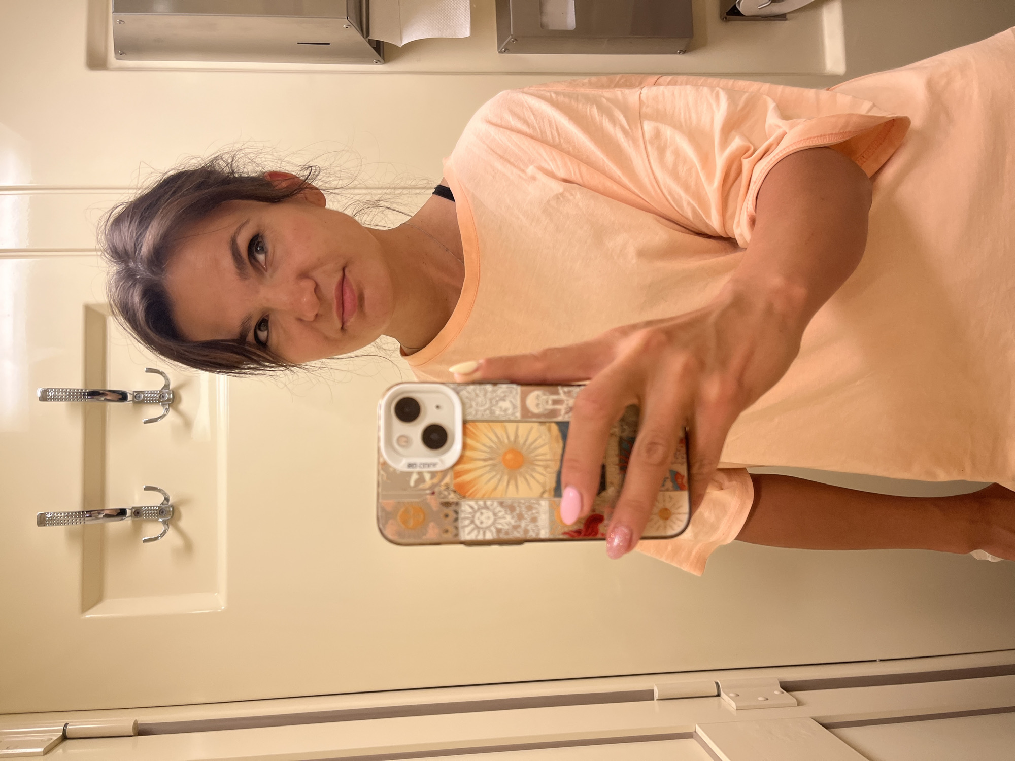 A woman with brown hair is taking a mirror selfie in what appears to be a small, enclosed space, possibly a train compartment or a bathroom. She is wearing a light peach-colored t-shirt. Her right hand holds a smartphone with a decorative case, and her nails are painted pink and glitter pink. She is looking slightly to her right with a neutral expression. In the background, there are two metal hooks mounted on a cream-colored wall, and a dispenser with white paper products on the right side. The overall lighting is soft and even. The location in the city of Лиски, Russia, is not specified in the image.