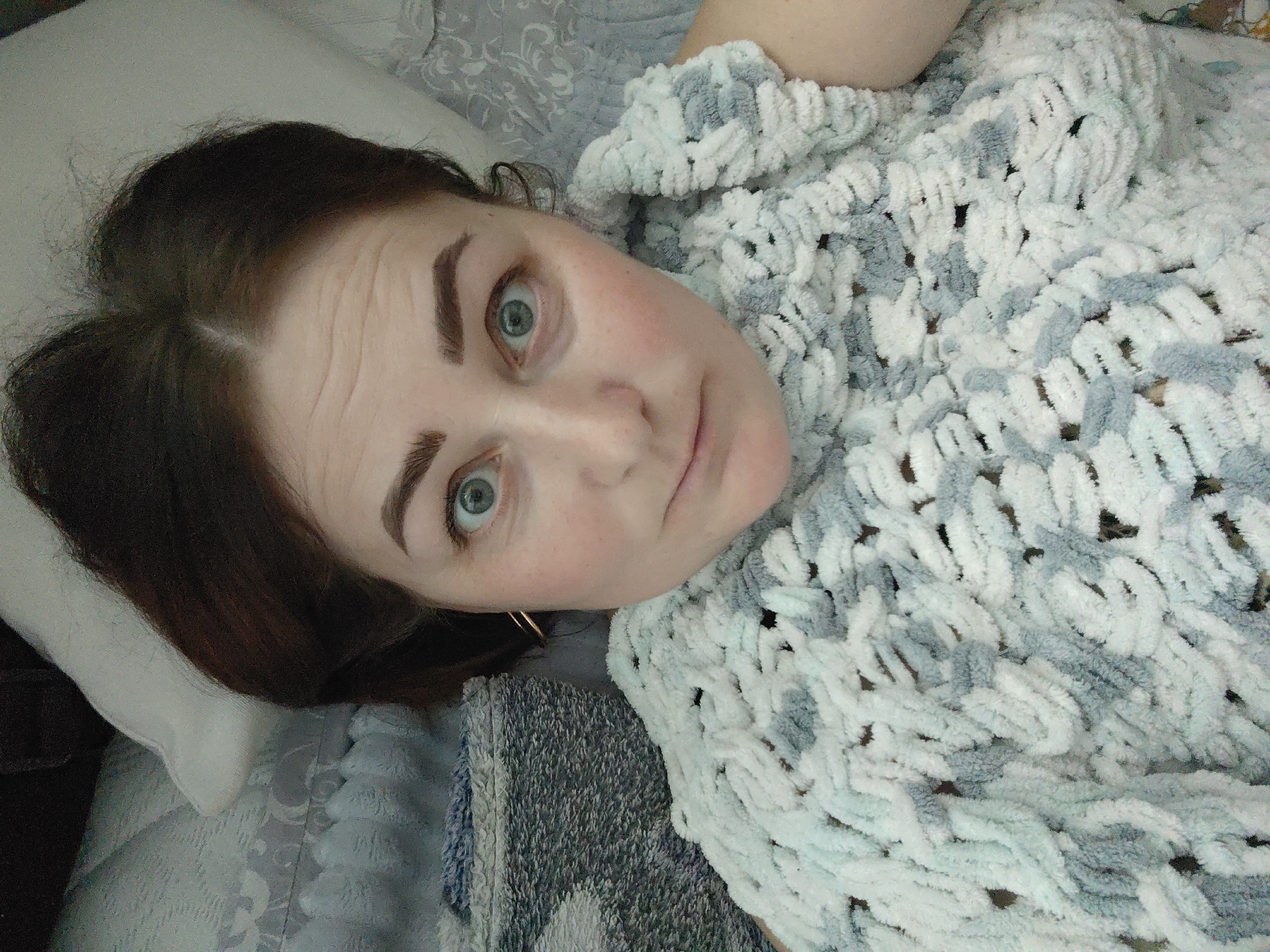 An individual, identified as an adult female, is depicted lying on a bed in an indoor setting in Markovskii, Russia. The subject has dark brown hair, distinct eyebrows, and blue-green eyes, which are widely open and directed towards the camera. Her skin tone is fair, with visible redness on the cheeks. Small, light-colored hoop earrings are present in her earlobes. A significant portion of her body is covered by a chunky knit blanket, patterned with alternating sections of light blue/teal and white yarn. Her head rests on a white pillow. The visible bedding beneath the subject and blanket features a light-colored fabric with a subtle floral or damask-like pattern. A darker, textured grey fabric is also visible in the lower left portion of the frame, adjacent to the main blanket.