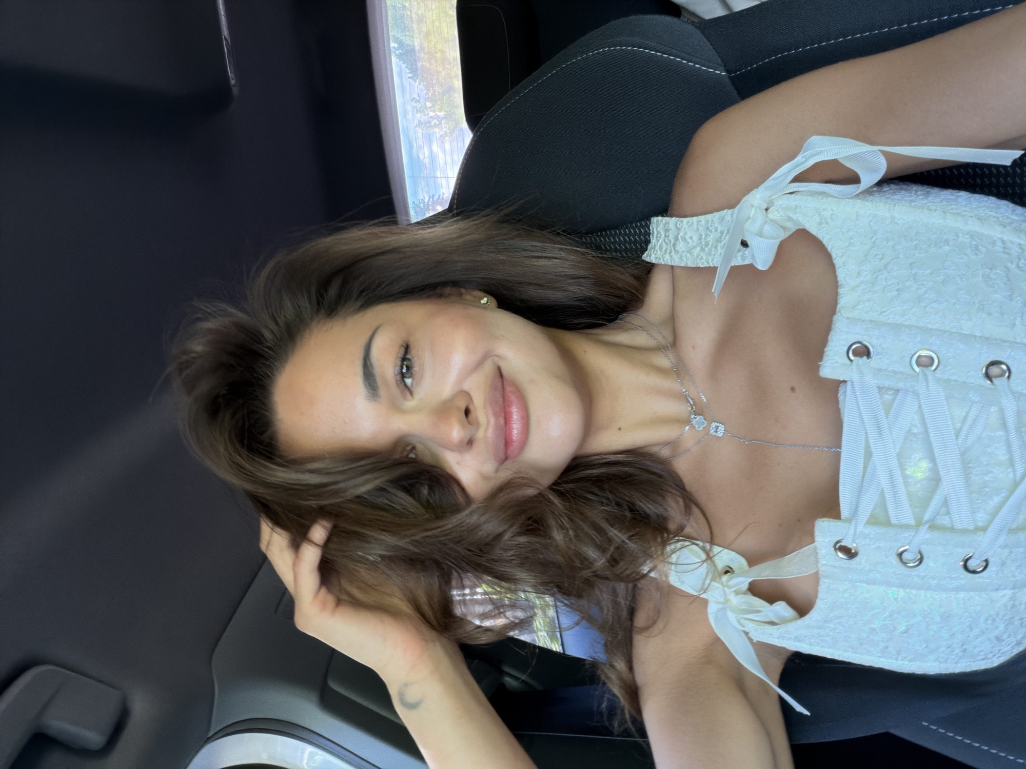 A woman with brown hair and fair skin is sitting in a car. She is wearing a white corset top and a silver necklace. Her left hand is in her hair. The background shows a car seat and a window with greenery outside. The location is Krasnoyarsk, Russia, based on the model's location.
