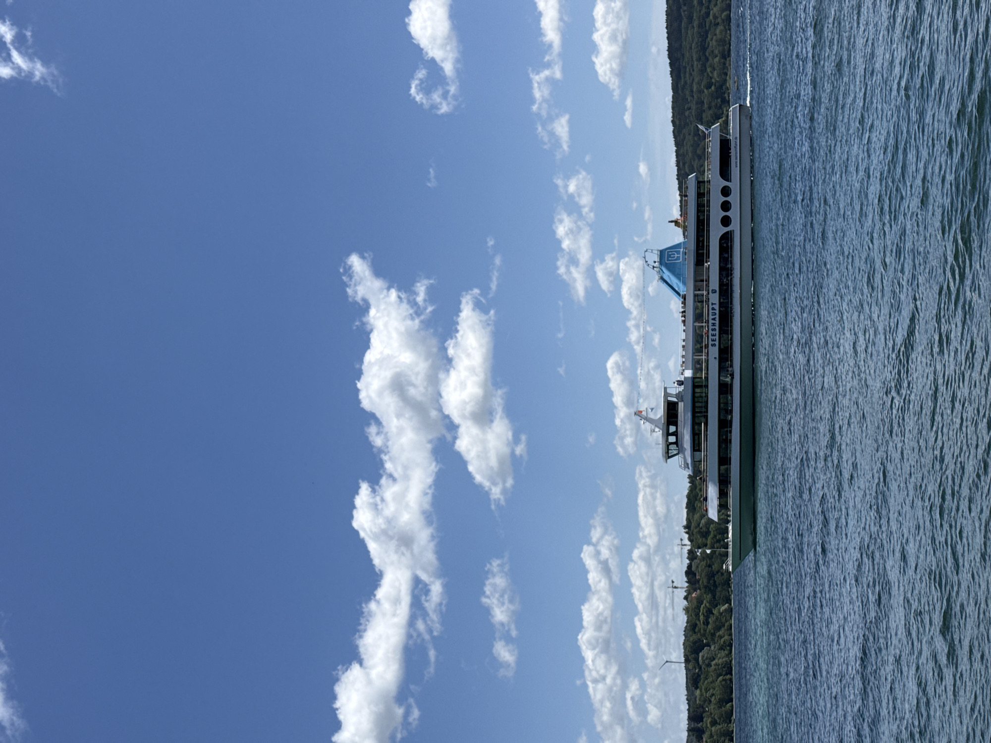 A wide shot shows a passenger boat traveling on a blue lake under a partly cloudy sky. The boat, named "SEESHAUPT," has multiple decks and is a light color with blue accents. Green, tree-covered hills form the shoreline in the background, with a wind turbine and construction cranes visible on the left. Several people are on the boat's decks. The water is choppy, indicating movement. The image appears to be taken from another boat or the shore.