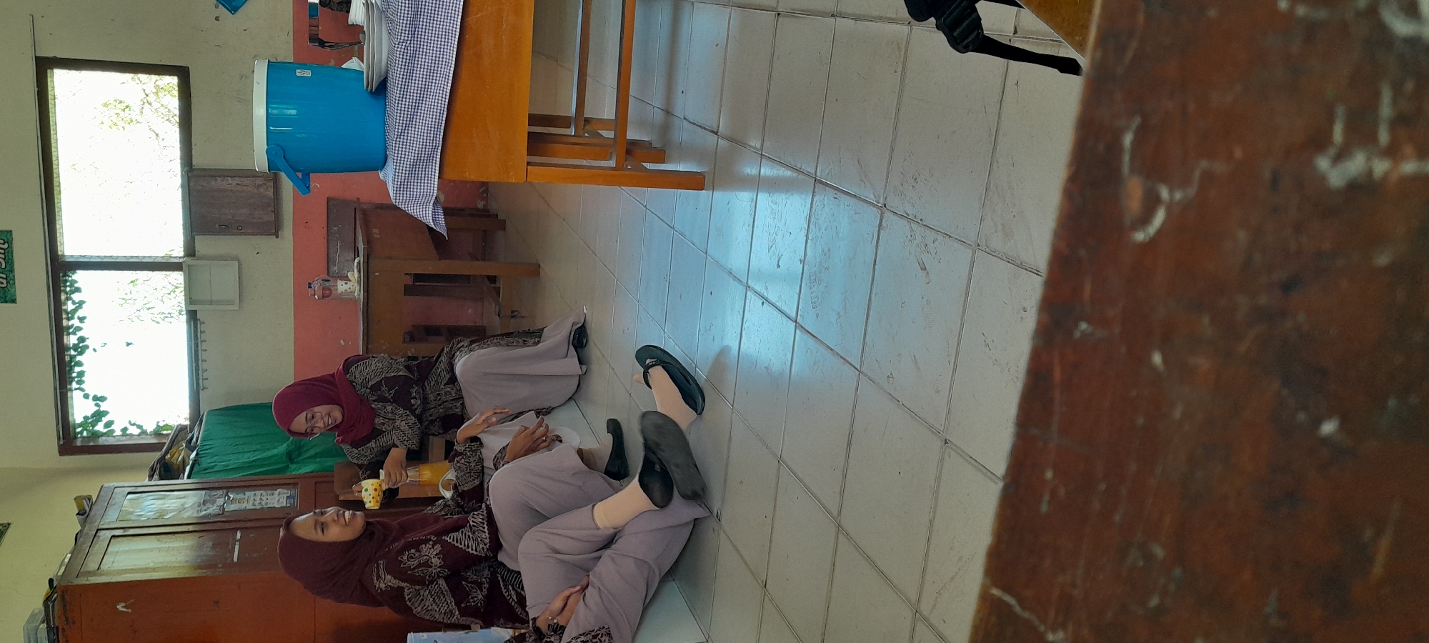 The image depicts an indoor scene, likely a classroom or a communal space, in Lemahbang Dewo, Indonesia. Two women wearing patterned maroon headscarves and matching uniforms are present. One woman on the left is seated, looking towards the other woman. The woman on the right, also seated, is pouring a beverage from a yellow pitcher into a cup. The environment features a light-colored tiled floor and a wall with a window that allows natural light to enter. Furniture includes wooden tables and cabinets. A large blue insulated container is visible on a table covered with a blue and white checkered cloth. The overall impression is a casual gathering.
