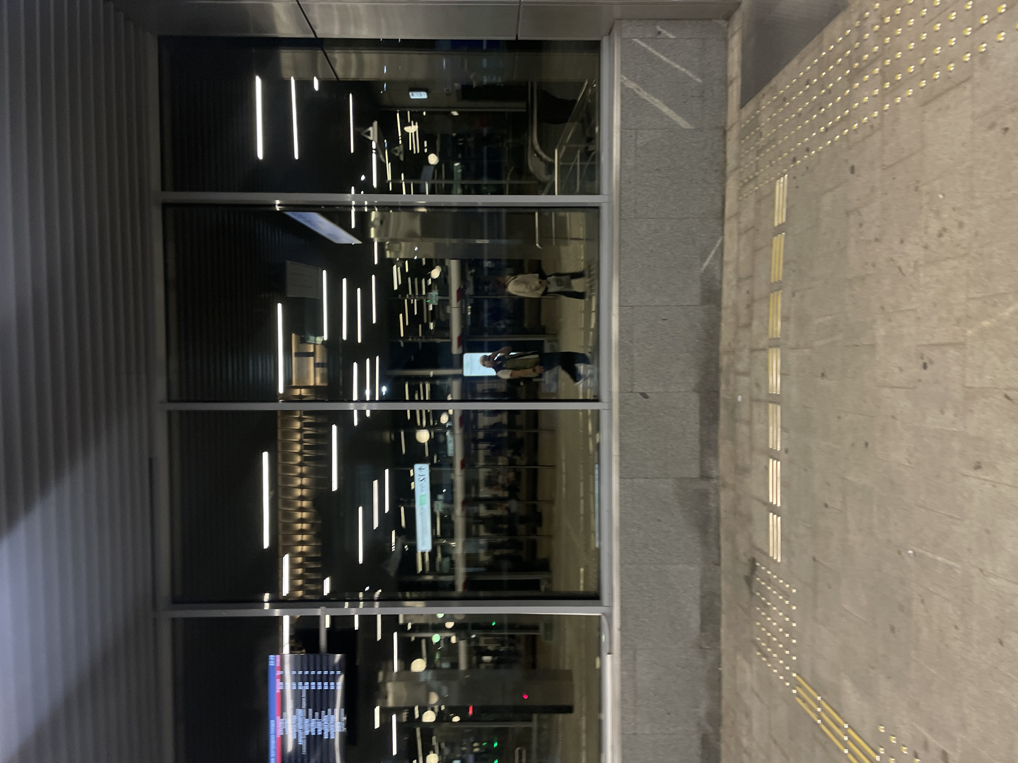 The image depicts a contemporary transportation hub, likely a metro station or train station, with large glass windows reflecting the interior. The scene appears to be in Moscow, Russia, as indicated by Cyrillic text visible on a digital display board in the upper left section of the image. The display shows departure and arrival times, with destinations like "МАЯКОВСКАЯ" (Mayakovskaya) and "ПЛ. РЕВОЛЮЦИИ" (Ploshchad Revolyutsii), which are well-known Moscow metro stations.
Inside the station, illuminated by horizontal fluorescent lighting, several people are visible. Two individuals are walking towards the right: one is wearing a light-colored jacket and carrying a bag, and another is wearing a dark jacket and a cap. A third person, in a green vest and cap, is also walking, partially obscured by a reflection. In the background, through the glass, more people and the platforms of the station are discernible. An escalator can be seen to the right, leading downwards.
The exterior foreground shows a tiled pavement with tactile paving strips, commonly found in public spaces to assist visually impaired individuals. The overall environment is clean and modern, suggesting an urban setting at night or in low light conditions. The reflections in the glass are prominent, blending the interior and exterior views into a complex visual composition.