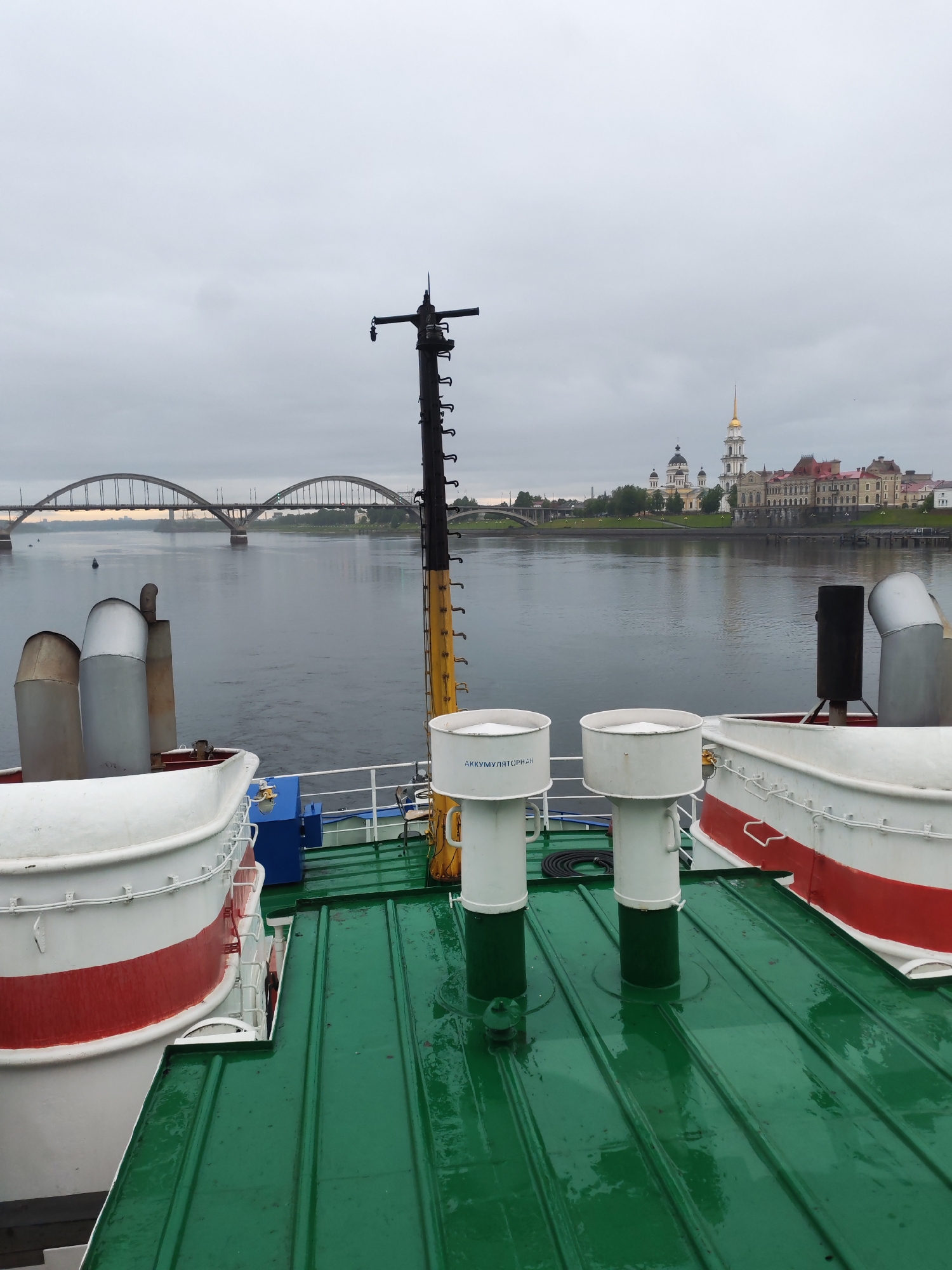 A medium shot, taken from the deck of a boat, shows a body of water leading to the city of Bol'shie Mkhi, Russia, which features a bridge and various buildings, including religious structures. The day is overcast with a gray sky.
The foreground is dominated by the green deck of the boat, streaked with moisture. Two white, cylindrical structures with green bases are prominent on the deck. These cylinders have inscriptions on them. The deck also features white and red safety barriers. In the midground, a black mast stands tall, with a yellow section near the base.
Across the water, a bridge is visible with two arched spans, leading to the city. The city is defined by various buildings, including structures with pointed roofs and a church or cathedral with a golden spire. The overall scene is one of quiet, scenic travel on a cloudy day.