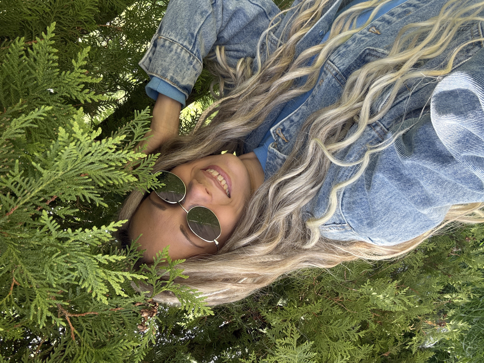 A woman with blonde wavy hair and round sunglasses smiles while lying among green coniferous plants. She is wearing a blue denim jacket over a blue sweatshirt. The location is likely outdoors, surrounded by evergreen foliage, but further specifics about Шебунино, Russia, are not discernible from the image.