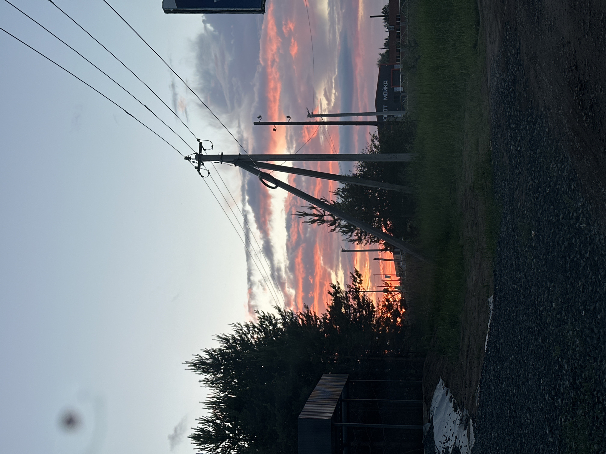 This image depicts a sunset scene on the outskirts of Chaikovsky, Russia. In the foreground, a gravelly area transitions into a grassy field. Silhouetted against the vibrant sunset sky are several power poles with wires stretching across the frame. To the left, a dark, rectangular structure, possibly a shed or part of a building, is visible. Further in the background, a building with signage that appears to read "РОБОТ МОЙКА" (Robot Car Wash) is present. The sky is filled with dramatic clouds illuminated by the setting sun, casting hues of orange and pink. The overall impression is one of a rural or semi-rural industrial edge of the city at dusk.