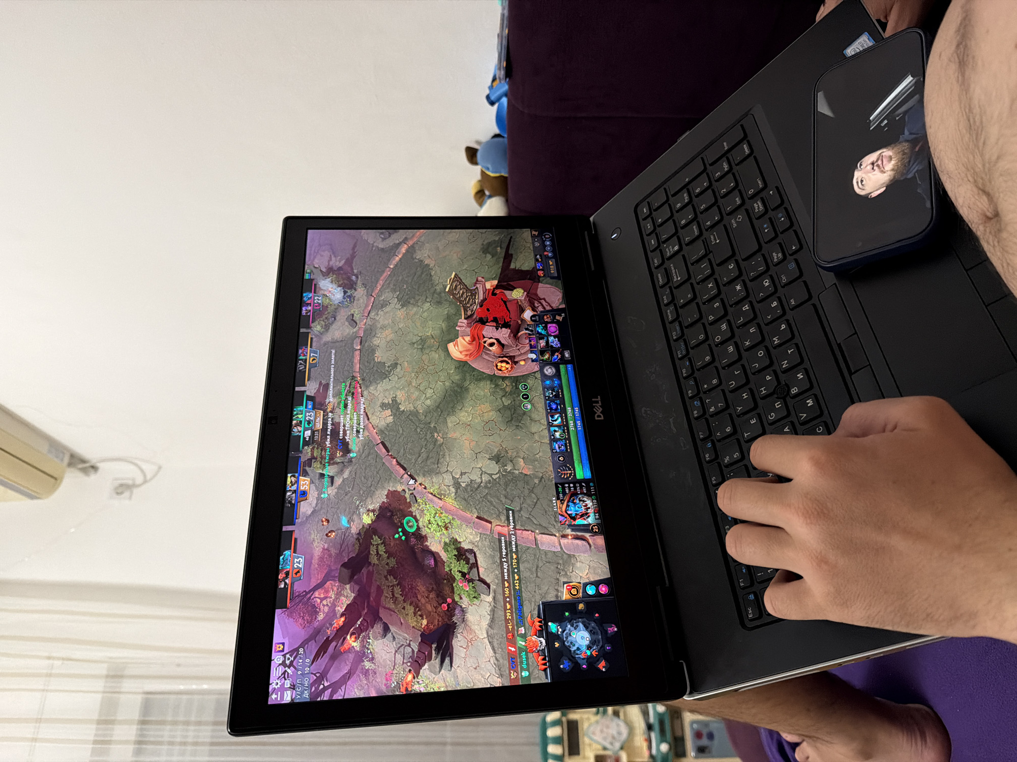A person is playing Dota 2 on a black Dell laptop. The laptop is sitting on the person's lap. A blue smartphone, showing the reflection of a man's face, sits on the laptop's keyboard. The person's hand is on the keyboard. The background includes a white wall, an air conditioning unit, and a purple couch with stuffed animals. Part of the floor is visible and has an indistinct pattern.
