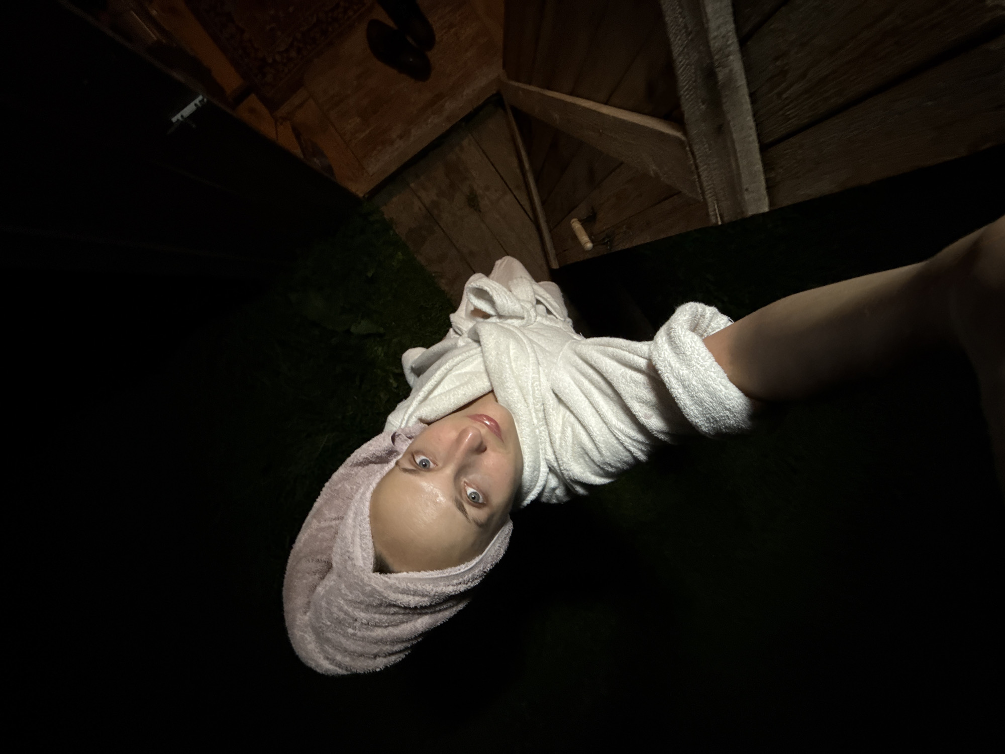A woman wearing a white bathrobe and a pink towel wrapped around her head is captured in a close-up selfie. She has light skin, blue eyes, and is looking directly at the camera with her lips slightly parted. The background is dark, with some wooden structures visible to the right, possibly part of an outdoor area or a shed. The lighting suggests it is nighttime. The image does not provide any specific details about the city of Sharkan, Russia, or any other location information.