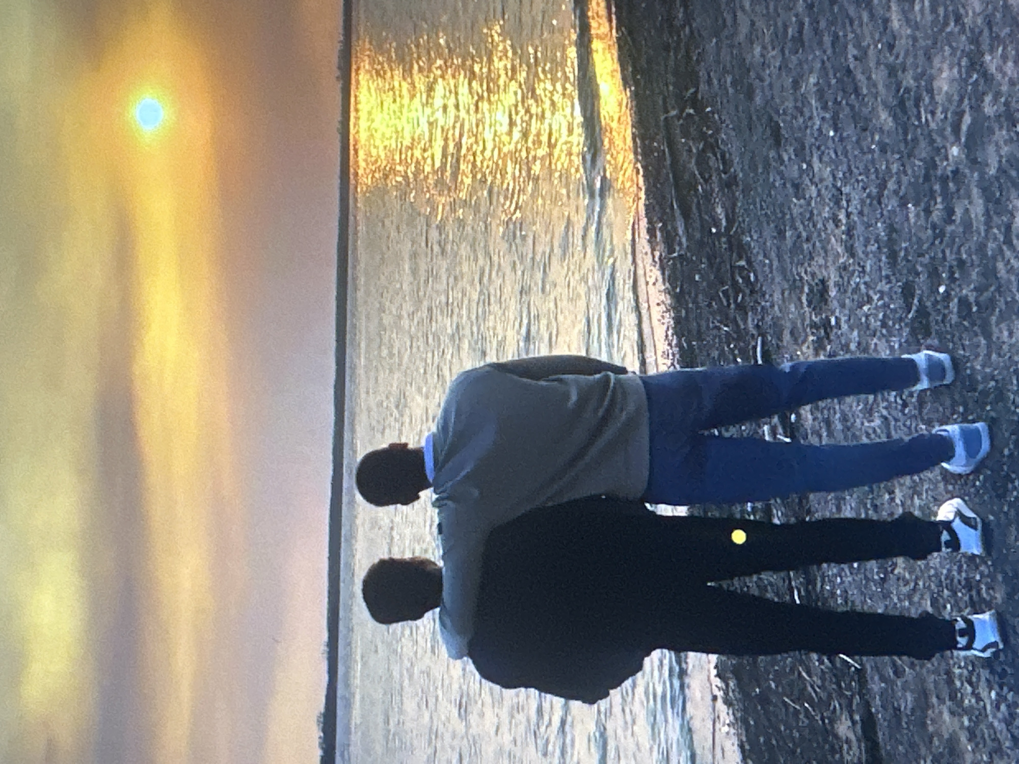 The image depicts two individuals standing on a dark, rocky shoreline, looking out at a body of water. The sky is dominated by a warm sunset, with hues of yellow, orange, and pink, and a bright sun or moon is visible in the upper right portion of the frame. The water reflects the vibrant colors of the sky, creating a shimmering effect on its surface. One individual has an arm draped around the other's shoulders, suggesting a close relationship. Both are dressed casually, with one wearing a dark top and black pants, and the other a light-colored sweater and blue pants. The shoreline is littered with small stones and debris. The overall scene is serene and atmospheric. The image is not taken in Tallinn, Estonia.