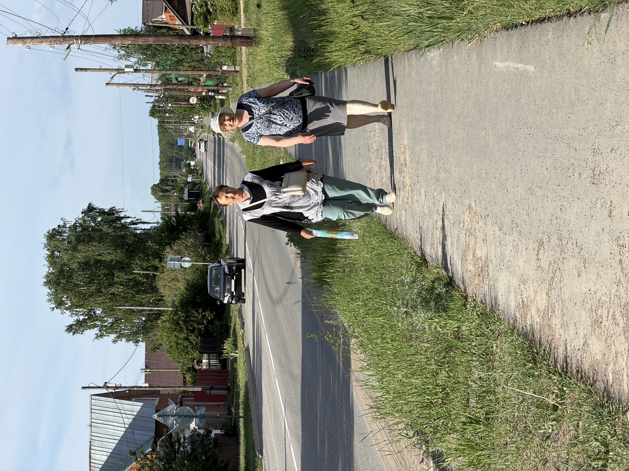 Two older women walk along a sidewalk next to a road in the city of Chaikovsky, Russia. Both women are wearing patterned blouses, light-colored purses, and light-colored shoes. The woman closer to the camera is wearing green pants and holding a colorful, folded object in her hand. The other woman is wearing a gray skirt and a light-colored hat. A black SUV is driving on the road. Utility poles line the side of the road. Houses are visible in the background. The sky is blue. Grass grows along the edge of the sidewalk.