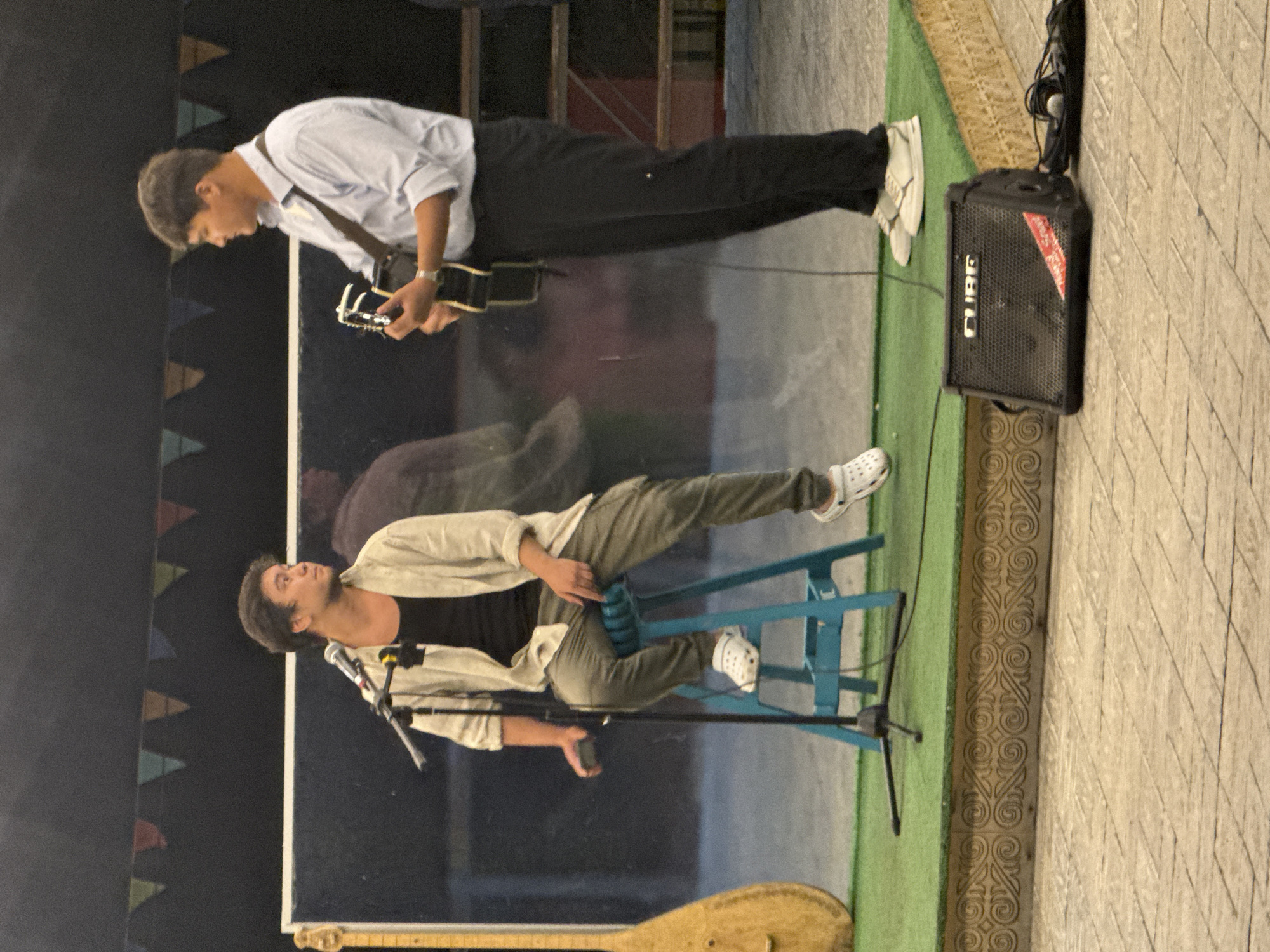 Two young men are performing music on an outdoor stage. The man on the left is seated on a blue stool, wearing a light-colored open shirt over a black tank top and grey pants with white Crocs. He is holding a microphone and looking to his right. To his left, a traditional stringed instrument with a long neck and a light-colored body is visible.
The man on the right is standing and playing an acoustic guitar. He is wearing a light blue collared shirt and dark pants with white sneakers. He is looking at the man on the left.
In front of the stage, there is a black "CUBE" brand amplifier and a microphone with its cable. The stage is covered with green Astroturf and has a decorative border. The background appears to be a dark wall with a row of colorful triangular flags.
The pavement in front of the stage is made of textured, light-colored pavers. The scene suggests a performance, possibly at a public event or cultural gathering. The presence of the traditional instrument could indicate a local or folk music performance. Given the context of Pavlodar, Kazakhstan, this could be a performance at a park, a street festival, or an outdoor concert venue.