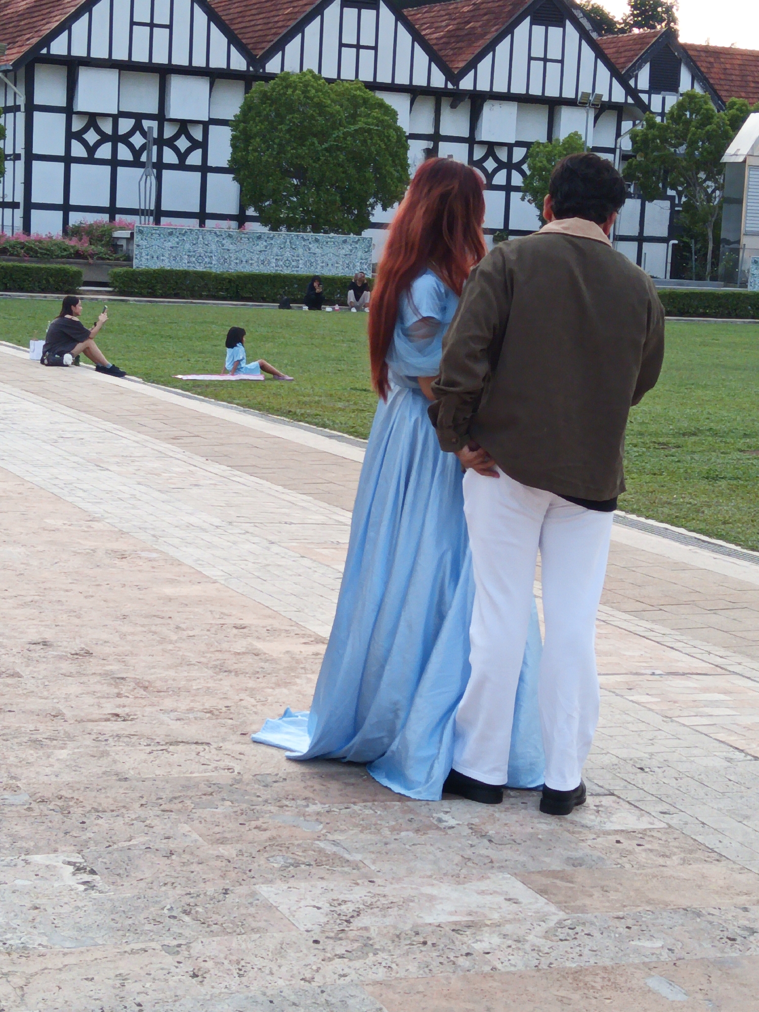 Two individuals are standing on a paved walkway in front of a grassy area with a building in the background. The person on the left has long, reddish-brown hair and is wearing a long, flowing light blue dress. The person on the right is wearing a dark olive green jacket and white pants. A group of people are seated on the grass in the middle ground, and another individual is seated further to the left. The building in the background has a distinctive black and white Tudor-style facade with a red tiled roof. The environment appears to be a park or a public outdoor space. This location is likely Dataran Merdeka, Kuala Lumpur, Malaysia, given the architectural style of the building which resembles the Sultan Abdul Samad Building.