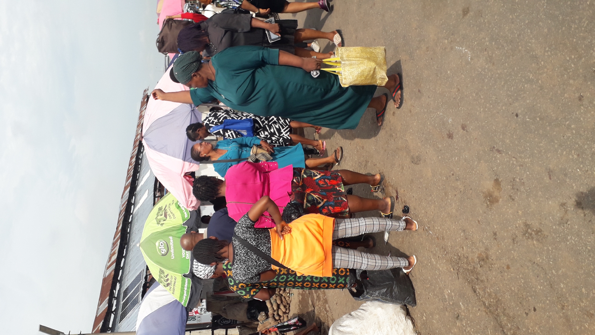 The image shows a group of people, mostly women, gathered in an outdoor market scene, possibly in Onitsha, Nigeria. The perspective is from above, looking down at the subjects. Several women are dressed in brightly colored clothing and some are carrying bags. One woman in a green dress holds a golden bag. There are also several umbrellas, one green and one pink and purple, providing shade. The ground appears to be a paved road or market floor. Some of the visible text on the green umbrella includes "glo Unlimited VOIC DAT" and "ONE CODE FOR EVERYTHING". The sky is visible in the upper left, suggesting it might be daytime. The overall impression is of a busy market environment.