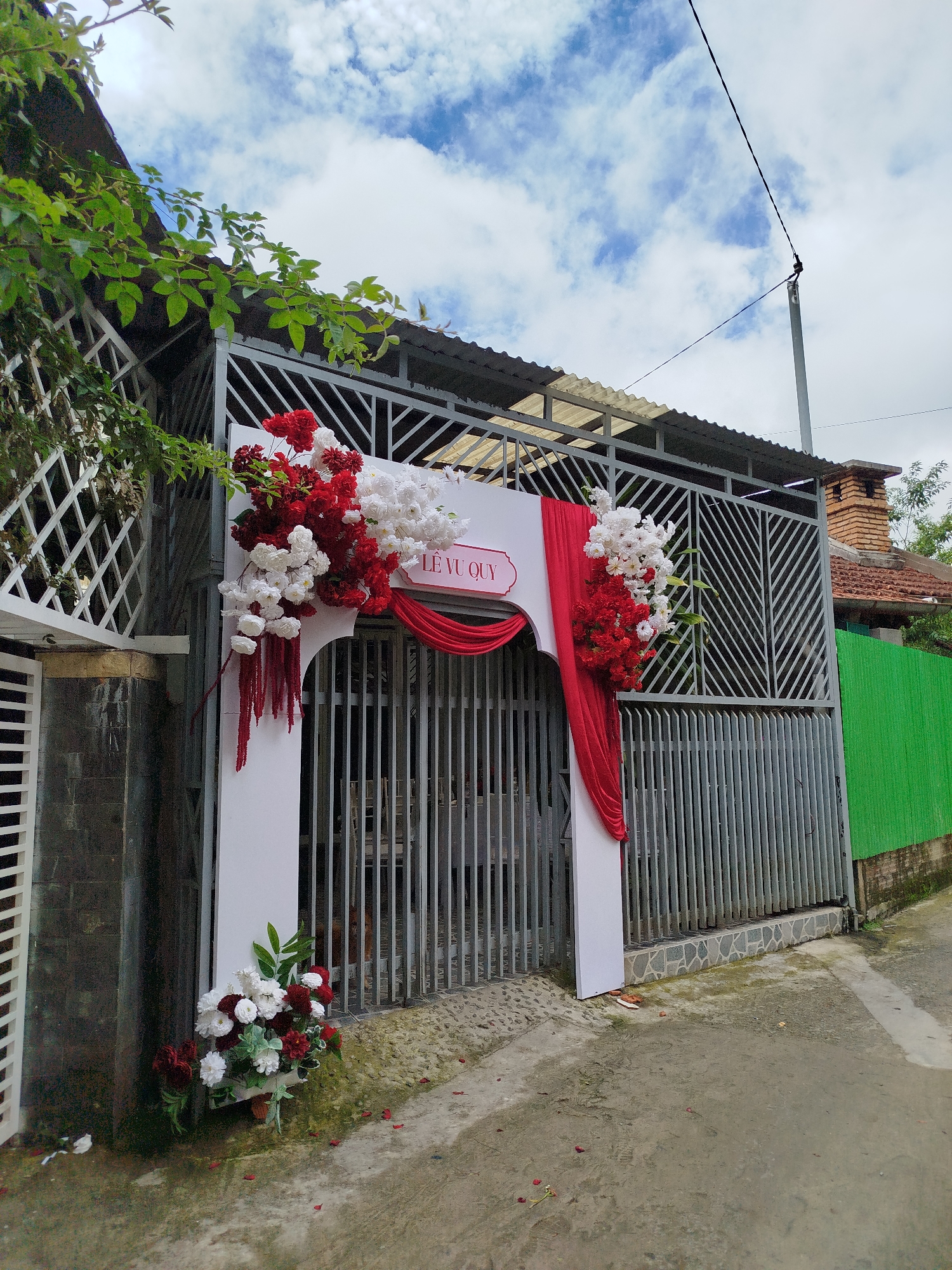 An outdoor scene in Da Lat, Vietnam, captures a decorated gateway, likely for a special celebration or ceremony. The main subject is a white, rectangular archway positioned in front of a grey metal gate, acting as an entrance. The arch is elaborately adorned with clusters of artificial red and white flowers, interspersed with green foliage. Red fabric is draped elegantly across the top and down the right side of the arch, complementing the floral arrangements. A smaller bouquet of red and white flowers sits at the base of the left arch support.
Above the arch, a white sign with a pink border displays the Vietnamese text "LÊ VU QUÝ," which translates to "Happy Wedding" or "Happy Marriage," indicating a wedding or engagement event.
The gate behind the arch is made of grey vertical metal bars with some diagonal patterns on the upper sections. To the left of the gate, there is a white lattice fence and lush green vines growing on a building structure. To the right, another section of the grey metal fence extends, and beyond it, a house with a reddish-brown tiled roof and a brick chimney is visible. A bright green corrugated metal fence separates the property from the street on the far right.
The sky is partly cloudy with patches of blue visible overhead. A utility pole with power lines is present in the upper right quadrant of the image. The ground in front of the gate is a concrete or paved surface, showing some moisture and a few scattered red petals, suggesting recent activity. The lighting indicates it is daytime. The vibrant decorations convey a festive and joyful atmosphere.