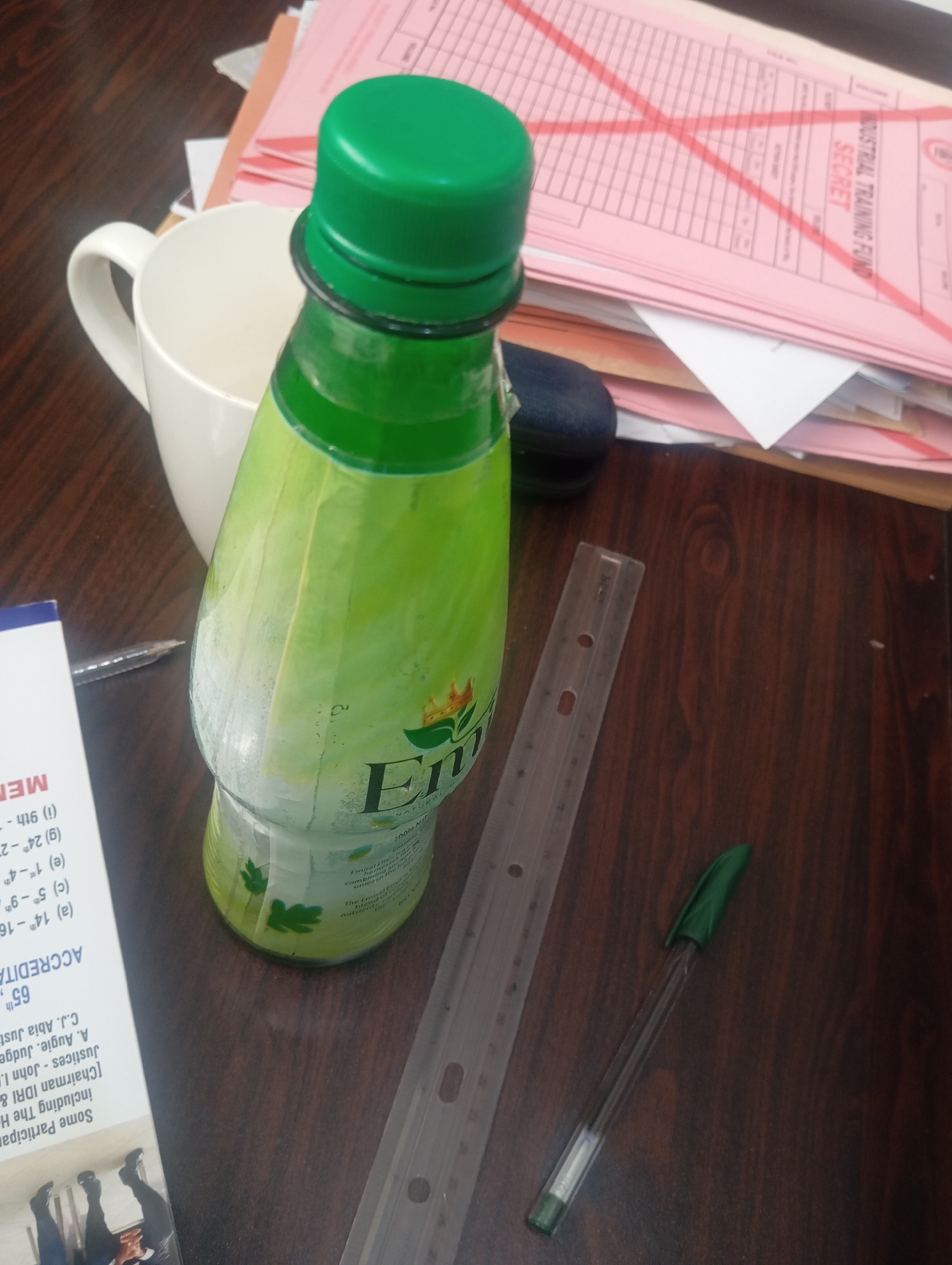 A bottle of "Emirates" brand drink with a green cap and label sits on a wooden desk. The bottle is partially obscured by a white mug on the left. In the background, stacks of pink and white papers are visible, one of which has a large red "X" drawn across it. To the right of the bottle, a clear plastic ruler and a green pen lie on the desk. A small portion of a white leaflet with text in black is visible to the left of the mug. The scene suggests an office or study environment. The location in Jos, Nigeria, is not discernible from the image itself.