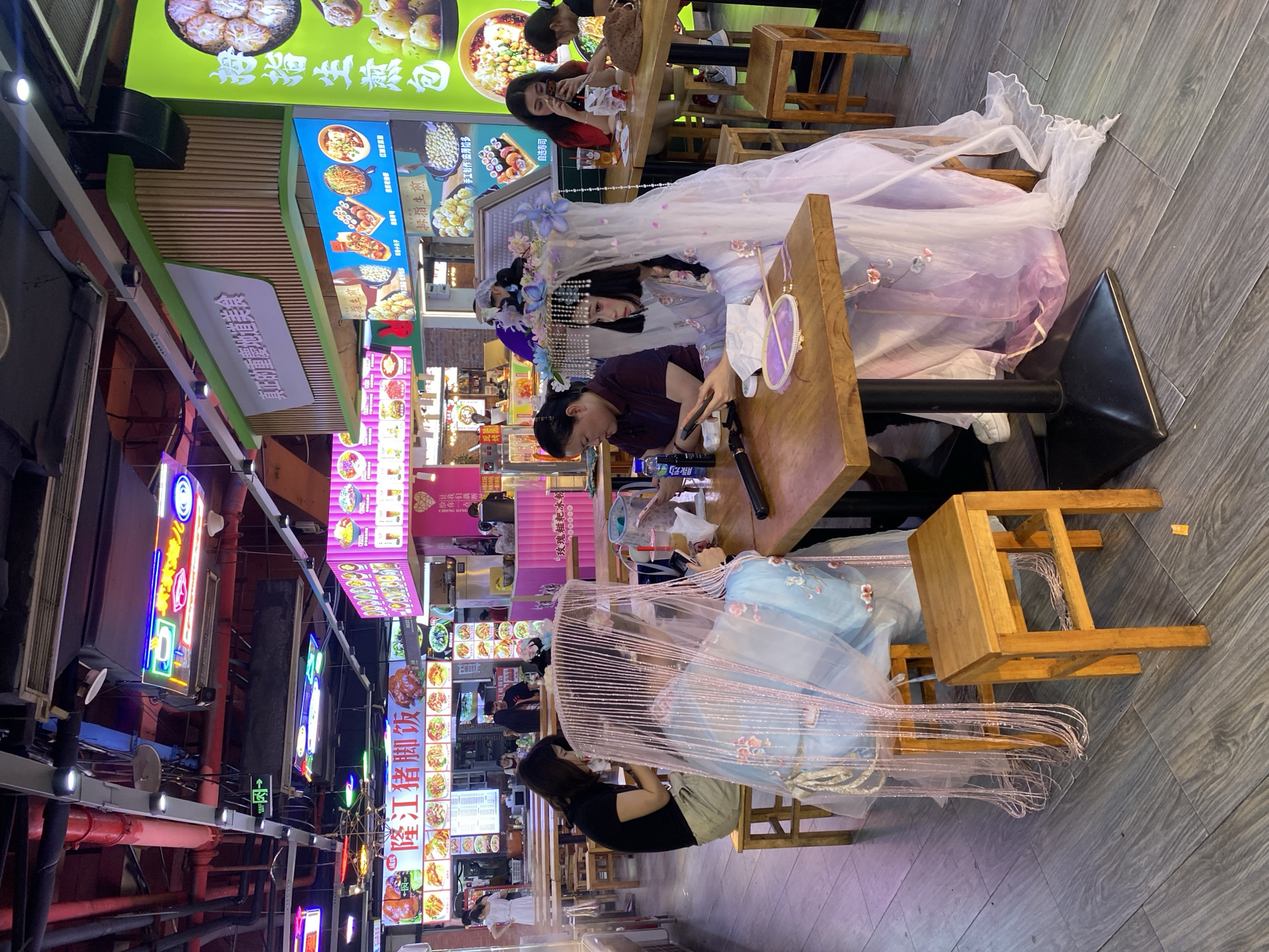The image depicts a bustling food court in Yuzhong District, China, with multiple food stalls and diners. In the foreground, a young woman dressed in elaborate traditional Chinese attire, complete with a headdress adorned with flowers and pearls, is seated at a wooden table. She is looking at her phone and holding a small fan. To her left, another young woman, also partially obscured by a sheer, beaded veil, is seated and looking at her phone. A third woman, dressed in everyday clothes, sits to the left of the veiled woman, also engaged with her phone. The table is set with a bowl of food and drinks. The food court environment is characterized by bright neon signs advertising various food items, overhead lighting, and numerous wooden tables and stools. In the background, other diners are visible, and more food stalls with menu displays can be seen. The overall impression is a lively and contemporary Asian dining scene.