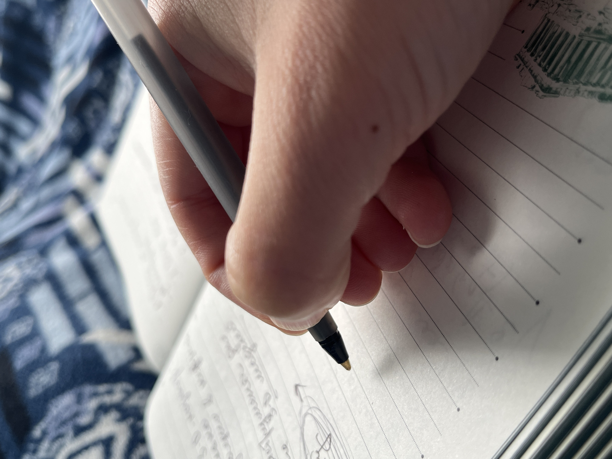 A close-up, vertical shot shows a hand holding a pen and writing in a notebook. The notebook has lined paper with small dotted lines. The pen is a clear plastic ballpoint with a grey barrel and a black tip. The hand is a light-skinned female's hand. The background is a blurry blue and white patterned blanket. The image is taken from a high angle, looking down at the notebook and hand. There are no visible indications of the location being in the city of ღრმაღელის თემი, Georgia.