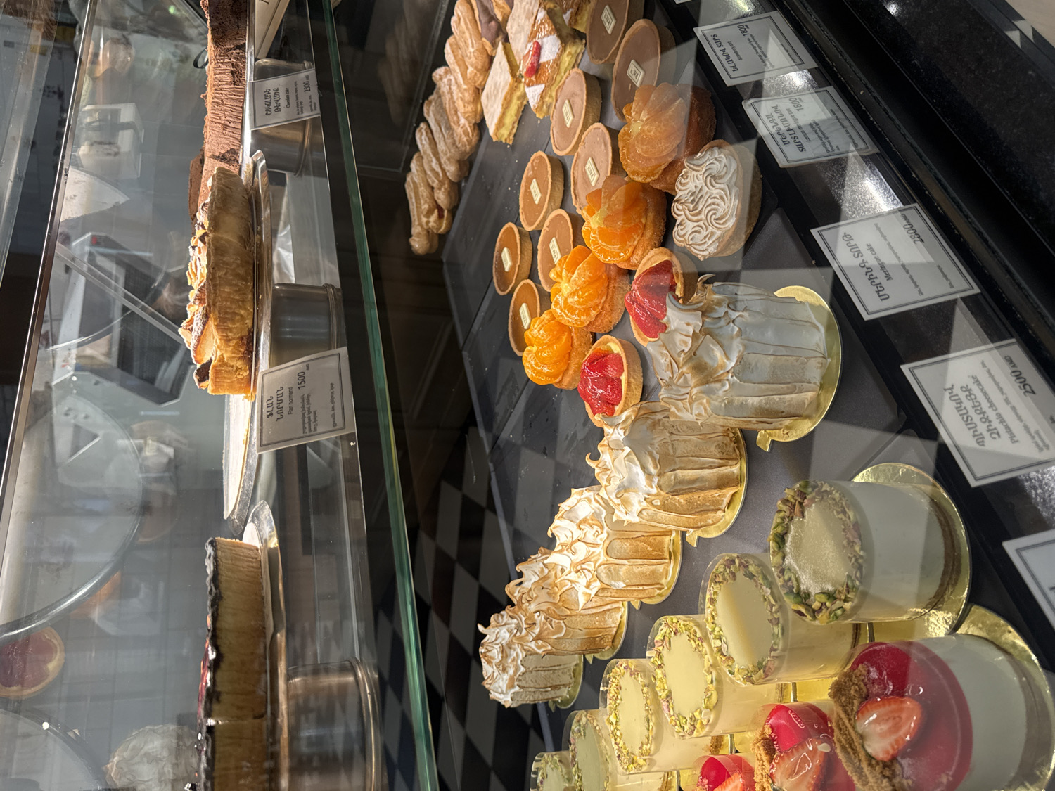 The image captures a vibrant display of various pastries and desserts inside a glass showcase at a bakery or patisserie in Yerevan, Armenia. The scene is brightly lit, highlighting the intricate details and colors of the confectionery.
The main subjects are numerous sweet treats, including flans, tarts, cheesecakes, and meringue-topped cakes. In the top left, a long, dark chocolate cake or roll, possibly a "Chocolate Roll," is visible. Below it, a "Flan normand" priced at 1500 AMD is displayed.
The central and right sections of the display showcase a variety of individual desserts. These include small tarts topped with vibrant orange segments (likely mandarins), tarts with red fruit fillings (such as "Strawberry tart" priced at 1800 AMD), and elegant cakes with piped meringue toppings, identified as "Meringue cake" (2300 AMD) and "Lemon meringue tart" (1800 AMD). Further along, individual "Pistachio cheesecake" portions, priced at 2500 AMD, are presented in small white cups with green pistachio rims. Some éclair-like pastries are also visible in the background.
Price tags are visible for most items, featuring both Armenian and English text, confirming the location context as Yerevan, Armenia, with prices listed in Armenian Dram (AMD). No people are visible, and the activity is simply the static display of baked goods for sale. The clean glass and organized presentation suggest a professional establishment. The time of day is not explicitly clear, but the artificial lighting indicates it could be during business hours.