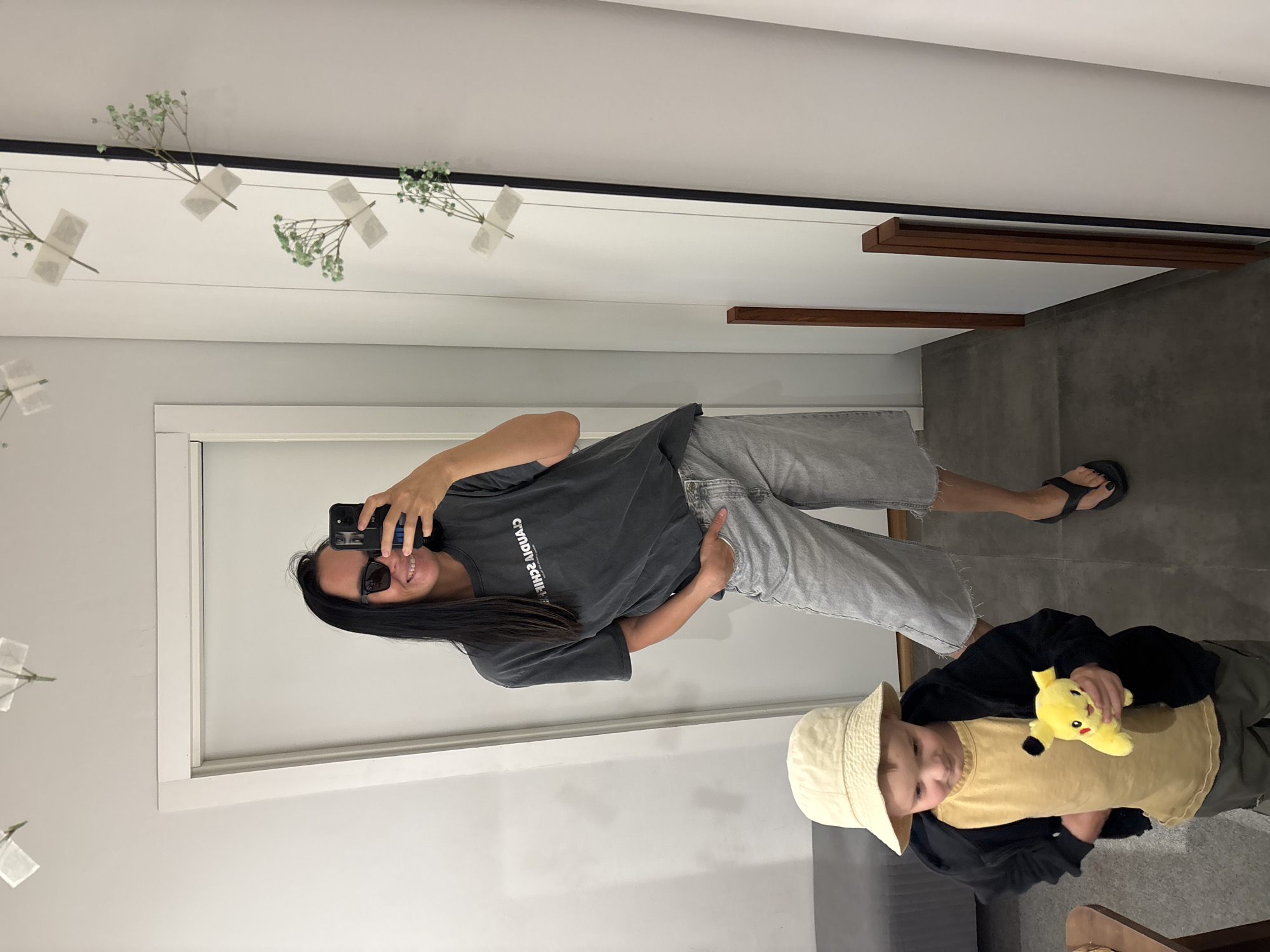 A person in a grey shirt and jeans is taking a selfie in a mirror. The mirror is mounted on a wall with white panels. There are decorative elements on the wall, including small taped flowers. A child is visible in the lower right of the frame wearing a hat, a yellow shirt, a black jacket and olive-colored pants. The child holds a Pikachu doll. The floor is tiled. The person wears sunglasses and sandals. The text on the shirt reads "STARDUST MACHINE". The location is Chaikovsky, Russia.