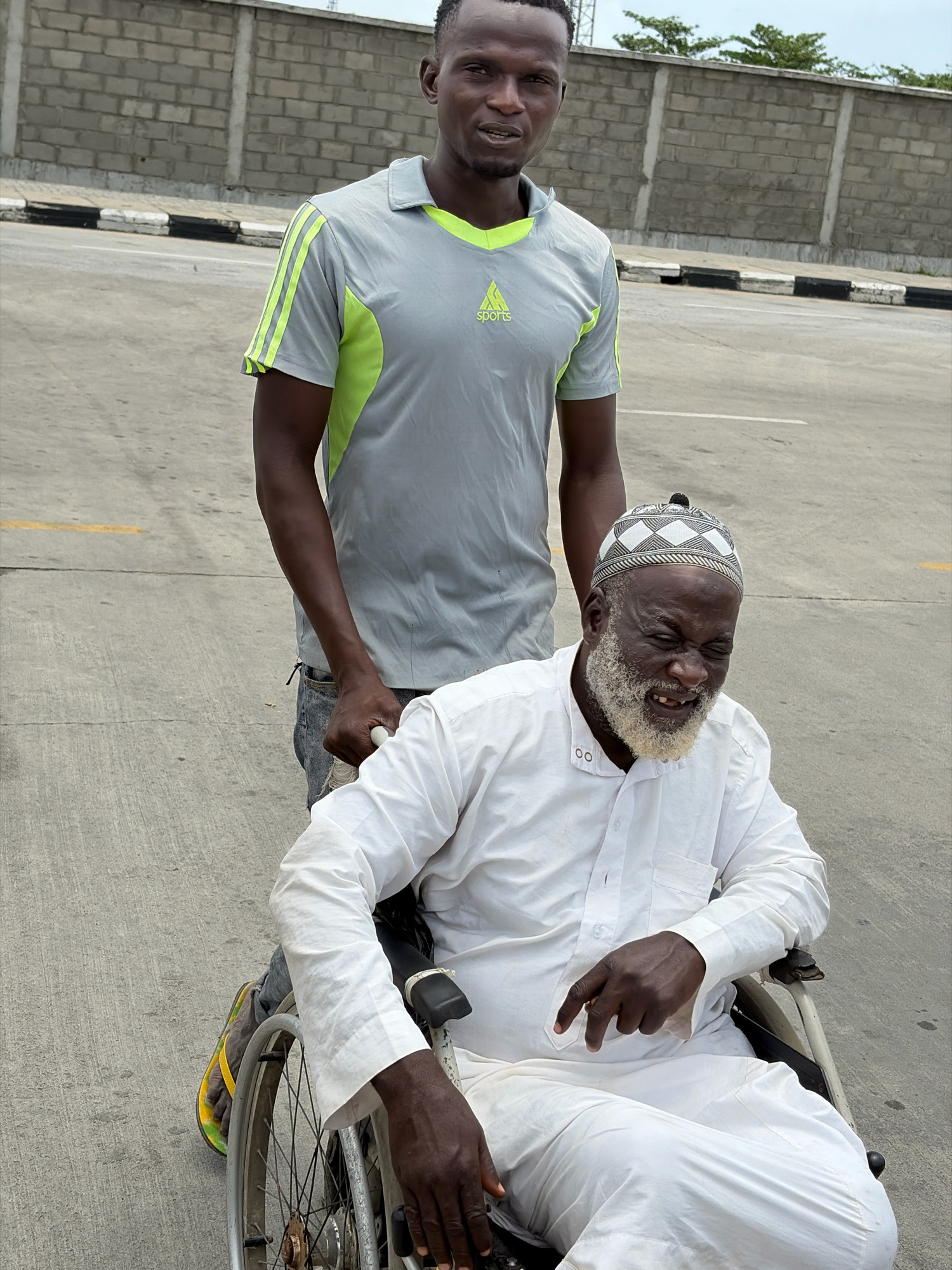 In the city of Jakande, Nigeria, a young Black man with dark skin and short hair, wearing a gray t-shirt with neon yellow accents and a pair of blue jeans, is pushing an elderly Black man in a wheelchair. The older man has dark skin, a gray beard, and is wearing a white traditional garment and a patterned cap. He is looking to his right with his eyes closed and mouth open as if laughing. The scene takes place outdoors on a paved surface, with a concrete wall and some greenery visible in the background. The overall lighting suggests it is daytime.