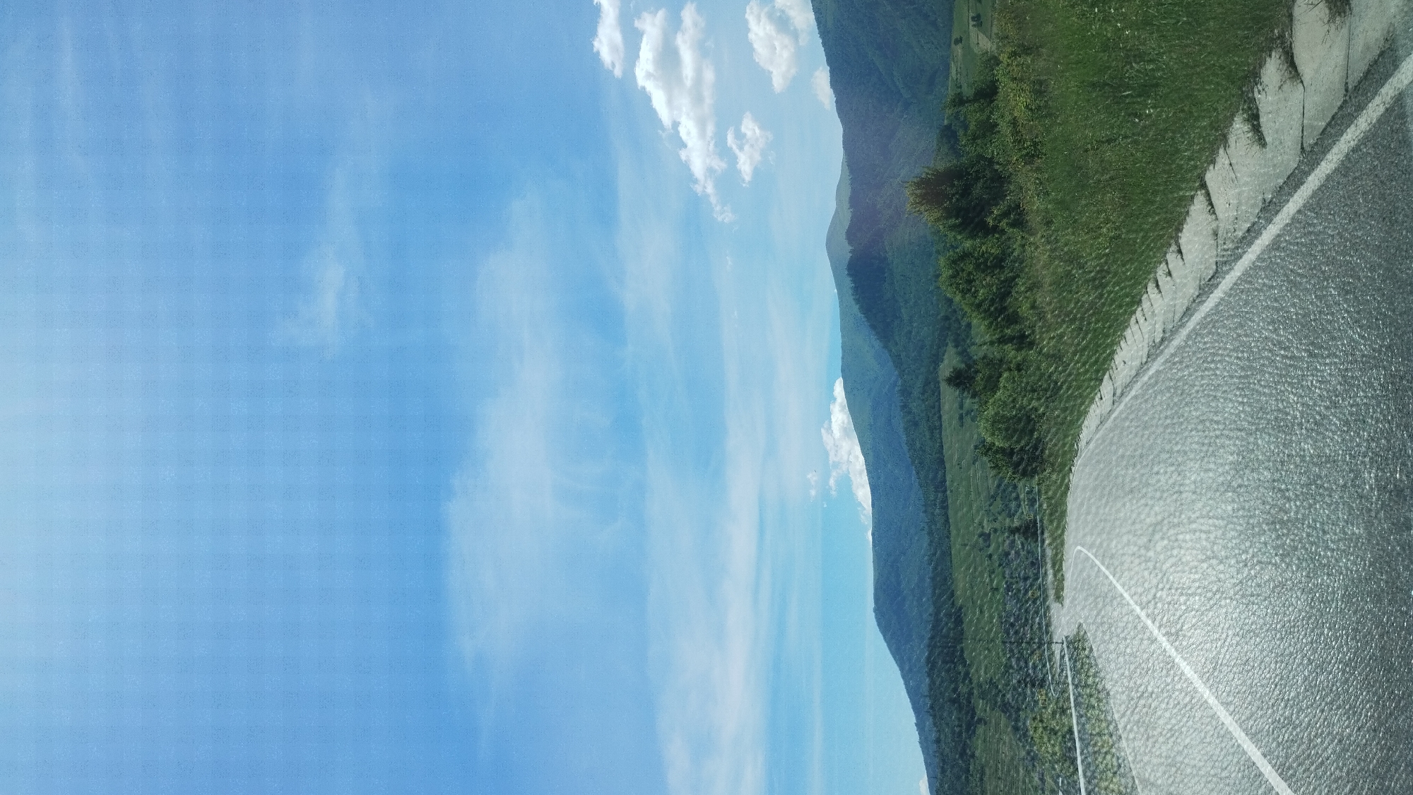 The image shows a paved road winding through a mountainous landscape on a bright, sunny day. The sky is a clear blue with scattered white clouds. The road has a white line marking the center. On the right side of the road, there is tall, green grass and a low concrete curb. In the background, there are rolling green hills covered in trees, with darker mountains in the distance. The overall scene suggests a rural or natural setting, possibly near Volovets, Ukraine, given the typical terrain of the Carpathian Mountains in that region. There are no people or specific objects clearly identifiable beyond the road, vegetation, and mountains.