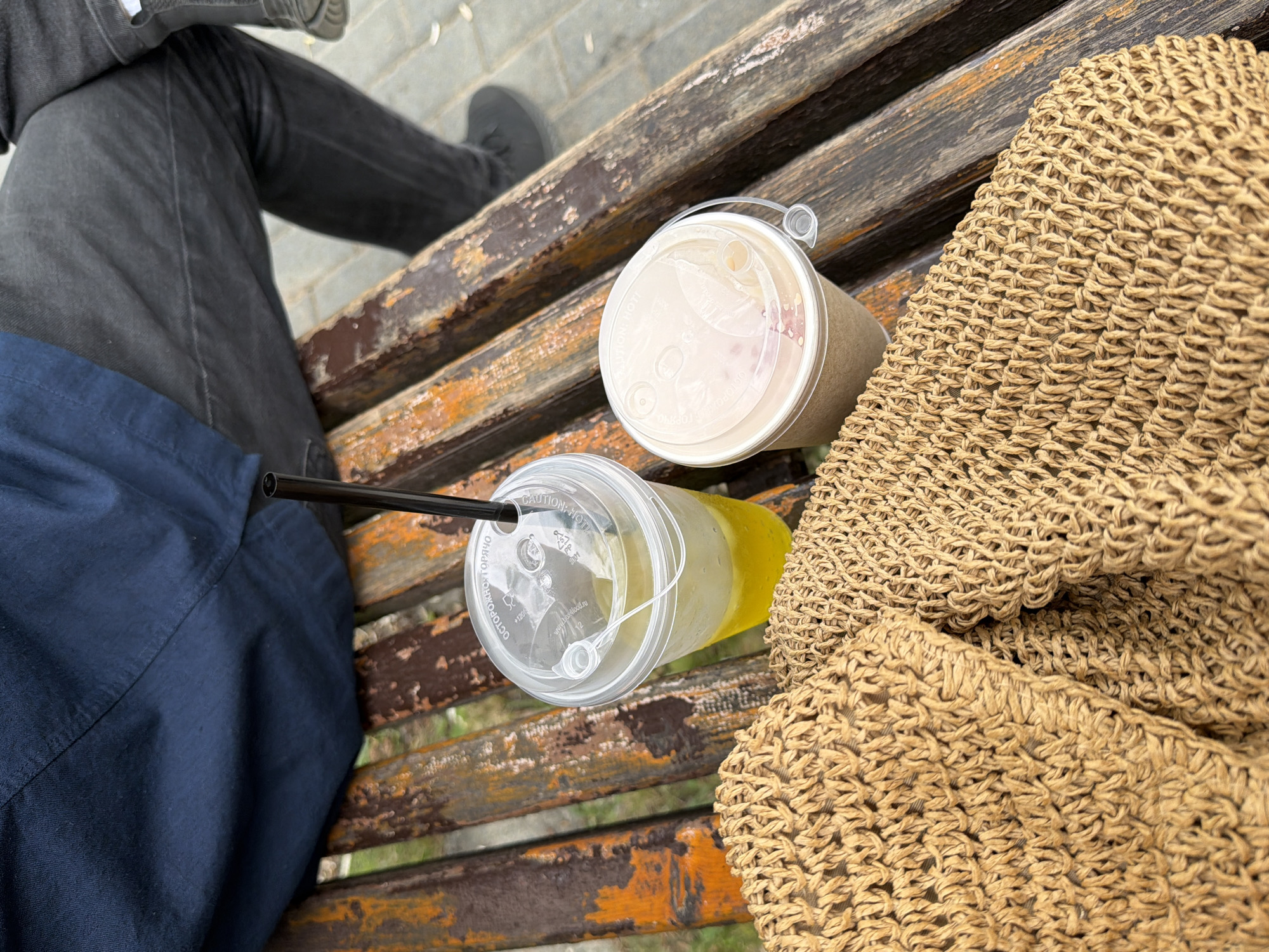 A person is sitting on a weathered, wooden park bench in Chaikovsky, Russia. The person is wearing a dark blue shirt and dark gray jeans. In front of the person, on the bench, are two drinks in plastic cups with lids and straws. One cup contains a yellow liquid, possibly lemonade, and the other contains a light-colored beverage, possibly coffee or tea, with pinkish-brown swirls visible. A woven straw bag rests on the bench in front of the drinks. The bench appears to be outdoors, with a paved area visible in the background, suggesting a public space like a park or promenade. The lighting indicates it is daytime.