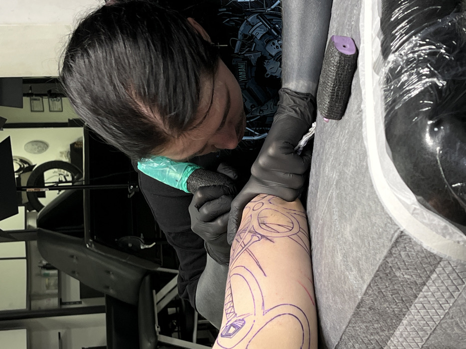 An individual, identified as a tattoo artist, is depicted meticulously tattooing a client's forearm within a professional studio environment in Rostov-on-Don, Russia. The artist, exhibiting dark hair, wears a black long-sleeved shirt and black disposable gloves. A light blue protective sleeve covers the tattoo machine in the artist's right hand, which is actively applying ink to the client's skin. The artist's left hand stabilizes the client's forearm, which features a complex tattoo stencil outlined in purple ink. The client's forearm rests on a grey, textured medical drape covering a surface. In the immediate foreground, a black textured grip wrap with a purple core lies on the drape, alongside another object partially covered in clear plastic wrap. The background is softly blurred but reveals typical tattoo studio furniture, including a black upholstered chair and shelving units. The scene emphasizes precision and sterile procedures within the tattoo artistry process.